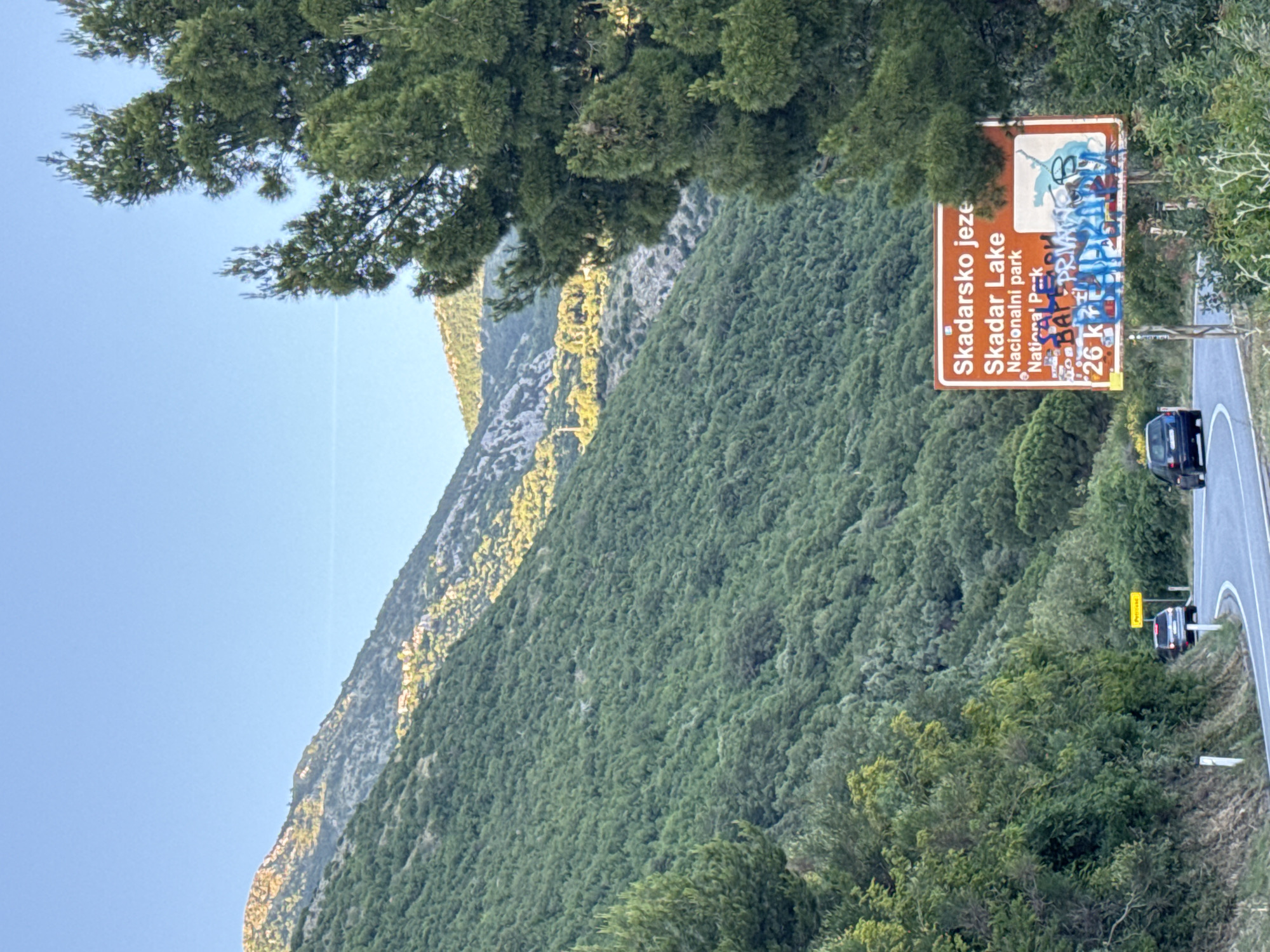 The image shows a mountain landscape in Medinski Krš, Montenegro, with a road winding along the side. A sign indicates Skadar Lake National Park, defaced with graffiti. Two vehicles are visible on the road. Dense vegetation covers the hillside.