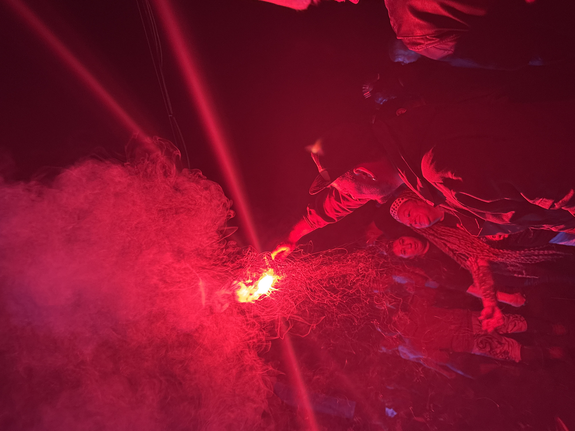 The image displays a nighttime scene in Большая Кивара, Russia, intensely illuminated by red light. A person, centrally positioned, wearing a dark cap and jacket, holds an activated pyrotechnic device aloft with their right hand. This device emits dense red smoke and bright yellow-orange sparks, which serve as the primary light source and pervade the atmosphere. To the right of the central figure, an individual with visible dreadlocks is partially depicted, appearing to smile. Multiple other human figures are present in the foreground and background, their features largely obscured by the pervasive red light, smoke, and shadows. One individual in the mid-ground left is visible wearing patterned shorts. Two distinct, parallel red light beams are evident in the upper right quadrant of the frame, cutting through the darkness. The overall setting appears to be an outdoor or large indoor space at night.