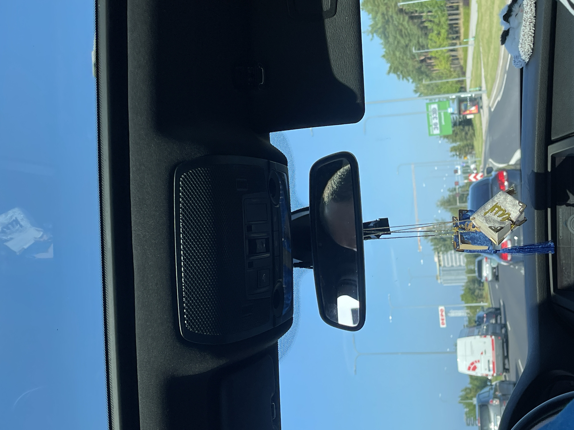 The image is taken from the perspective of a passenger inside a car, looking forward through the windshield. The view captures a section of a road in Tallinn, Estonia, under a clear blue sky. The car's interior is visible in the foreground, with the dashboard, steering wheel, and rearview mirror. A religious ornament hangs from the rearview mirror.
On the road, several vehicles are present, including a white van with red markings, and various cars. Traffic appears to be moving, indicated by the presence of vehicles in multiple lanes and some distance between them. Streetlights line the sides of the road, and on the right, there is a line of trees.
In the background, buildings and construction cranes are visible, suggesting an urban or suburban environment. A green sign with the number "36 339" is prominently displayed on the right side of the road. The overall scene depicts a typical day of driving in a city.