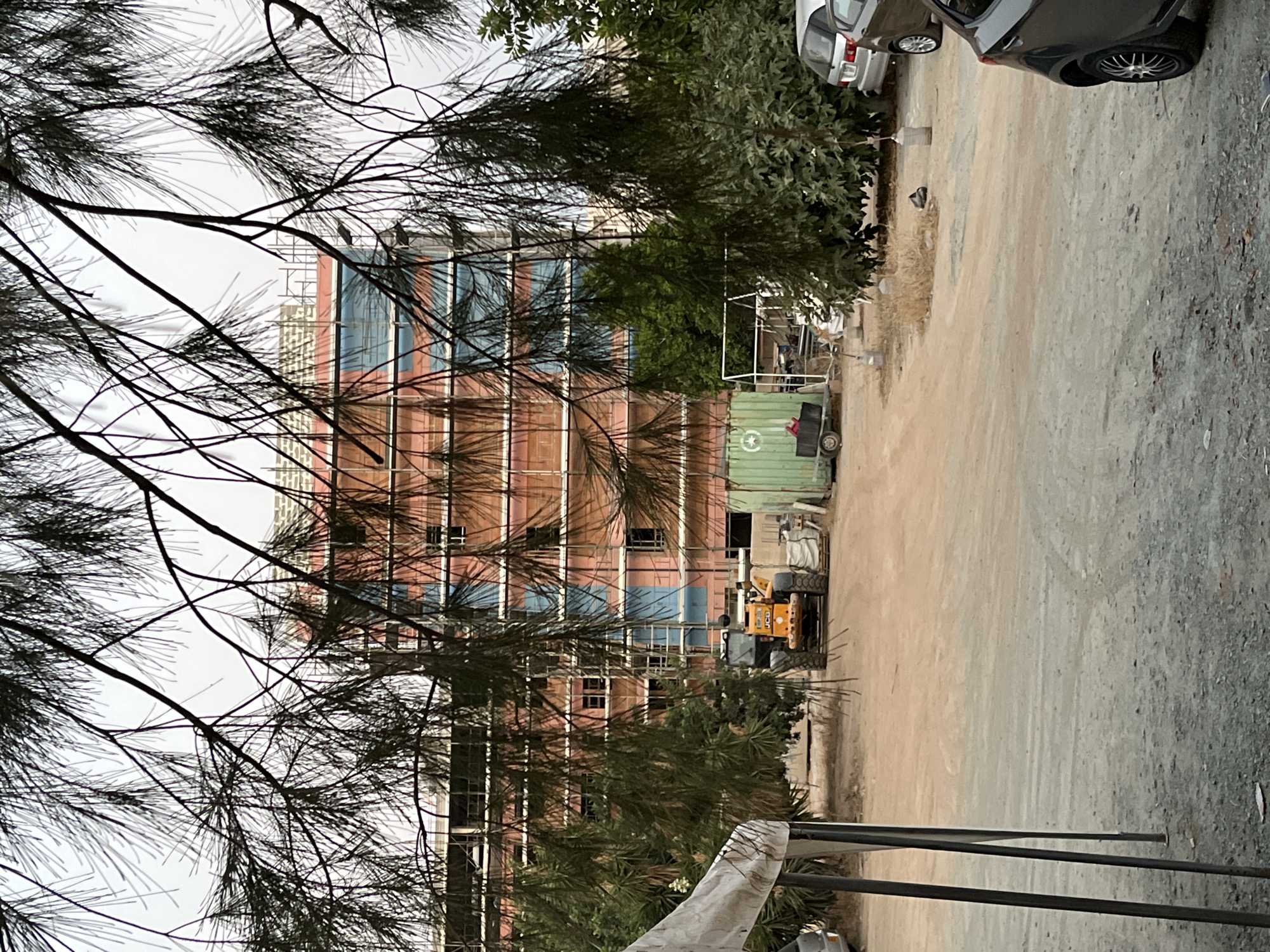 The image depicts a construction site in Limassol, Cyprus, as evidenced by the visible scaffolding and building materials. In the foreground, a yellow JCB loader is parked on a dirt surface, facing towards the right. To its left, a white tent structure is partially visible. Behind the loader, a green shipping container with a white circular logo is situated on a trailer.
The primary subject is a multi-story building under construction. It features exposed brickwork and blue sheeting covering sections of the facade, indicating ongoing work. The building is surrounded by scaffolding, typical for a construction project.
Several trees with sparse foliage are scattered throughout the scene, some closer to the foreground and others nearer to the building. The ground is a mix of dirt and gravel. In the background, two cars are parked on the right side of the image, one silver and the other dark grey. The sky is overcast, suggesting a cloudy day. The overall impression is one of an active construction zone in an urban or semi-urban setting within Limassol. No individuals are discernible in the image.
