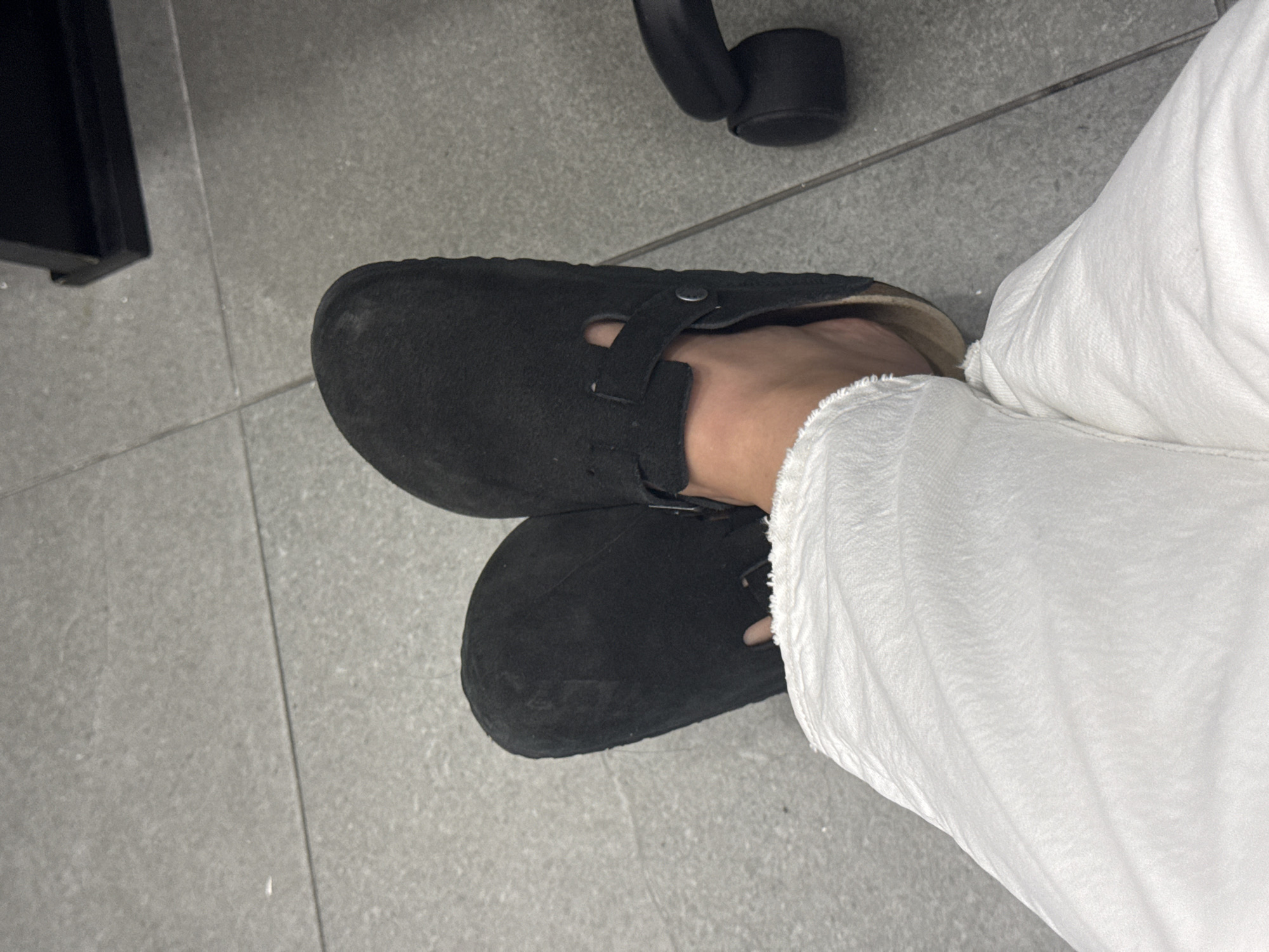 The image shows a person wearing a pair of black suede Birkenstock Boston clogs and white pants, likely sitting on an office chair. The setting appears to be an office with gray tiled flooring. There is no information to suggest this photo was taken in San Bernardino Tlaxcalancingo, Mexico.