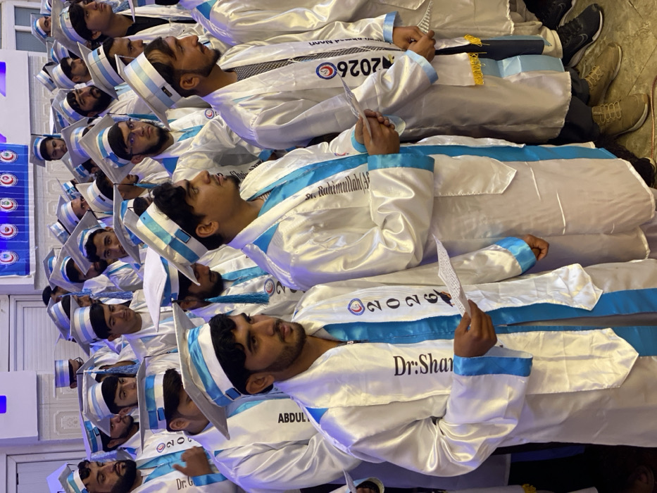 A group of men wearing white graduation gowns with blue accents and matching mortarboards are closely packed together. The gowns have names printed on them, such as "Dr.Shar" and "Abdul". The mortarboards have a white tassel and blue stripes. Each graduate holds a piece of paper, possibly a diploma or program. The photo is rotated 90 degrees, with the heads of the graduates on the left.