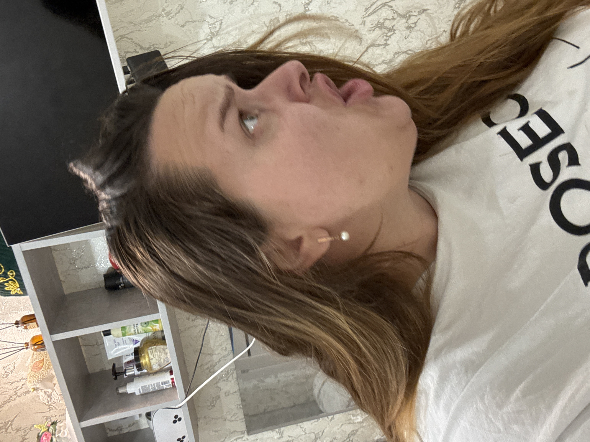 A medium-shot, profile view shows a young woman with long, brown hair, wearing a white t-shirt with black lettering. She is looking to her right with her mouth open in an expression of surprise or dismay. Her right eye is visible, and she is wearing a small, pearl earring in her right ear. Behind her, a shelf is visible with various bottles and jars, likely toiletries. The background is a patterned wallpaper. The location is noted as Dobrovske rural settlement, Ukraine.