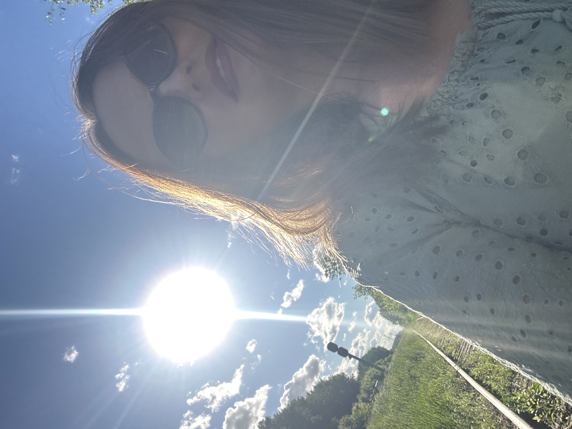 A woman wearing sunglasses and a light blue top stands on a railway track under a bright sun. The sun is positioned in the upper left quadrant of the image, emitting a strong glare that affects visibility. The woman is looking towards the camera, with her face partially obscured by sunglasses. The railway track runs diagonally from the lower left to the upper right of the frame, bordered by green grass and trees on either side. A signal pole is visible in the distance along the tracks. The sky is blue with scattered clouds. The scene appears to be outdoors, possibly in a rural or semi-rural area. The location of Medvedevo, Russia is not depicted or implied in the image.