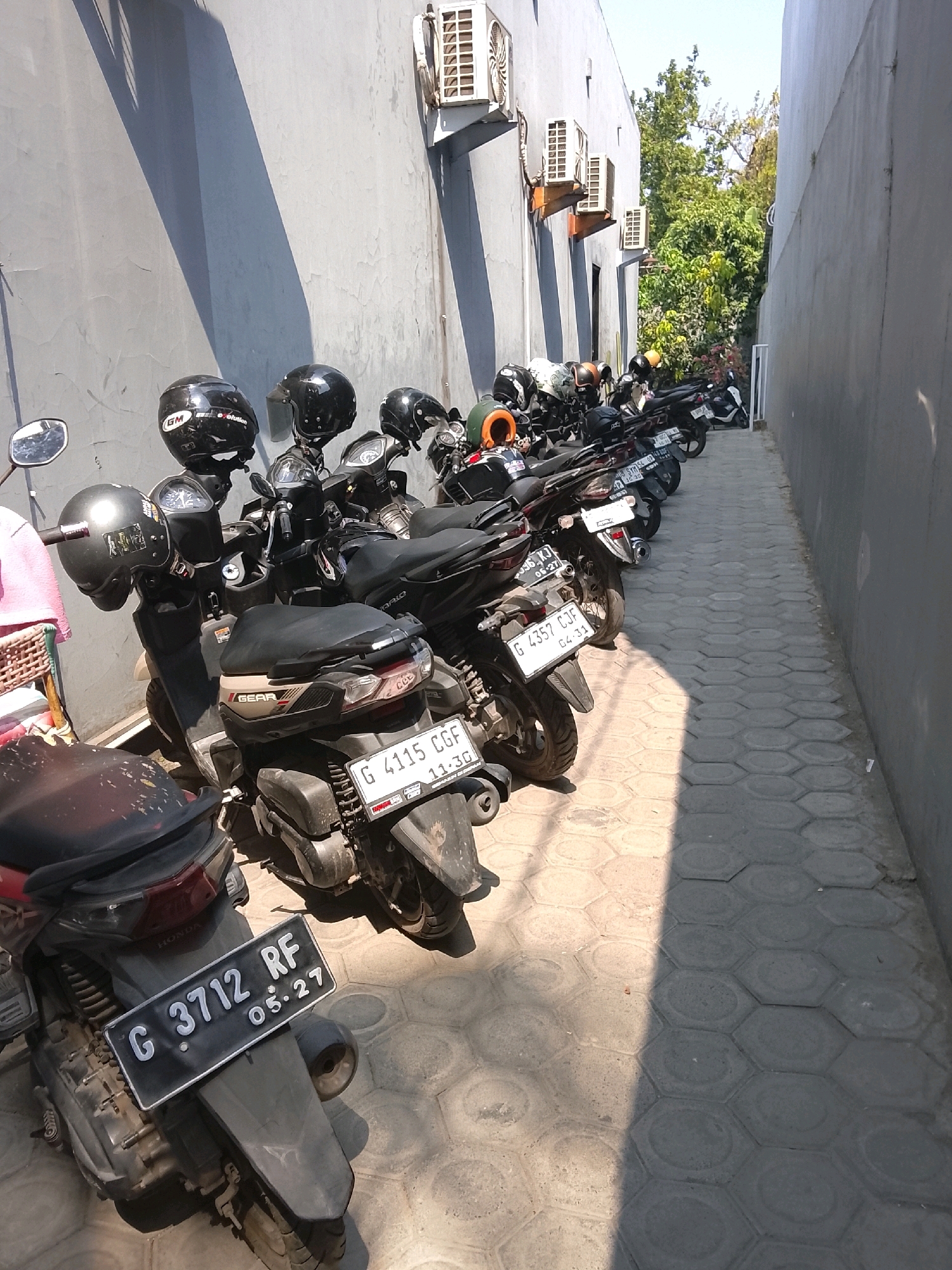 The image shows a narrow alleyway in Slawi, Indonesia, with several parked motorcycles. The alley is bordered by two buildings, one with a gray, textured wall and the other a lighter colored building with air conditioning units visible. The ground is paved with hexagonal interlocking concrete blocks, with a distinct shadow cast across the pavement from the left side of the image.
The motorcycles are mostly black, with various helmets placed on them. Some of the license plates are partially visible, including "G 3712 RF 05.27", "G 4115 CGF 11.30", and "G 4357 CJF 04.31". One motorcycle in the foreground has a license plate that reads "G 3712 RF 05.27". The motorcycles appear to be parked in a lot or on a street outside of a building. The overall scene is quiet and static, with no people or activity visible. The lighting suggests it is daytime, with sunlight illuminating parts of the alleyway.