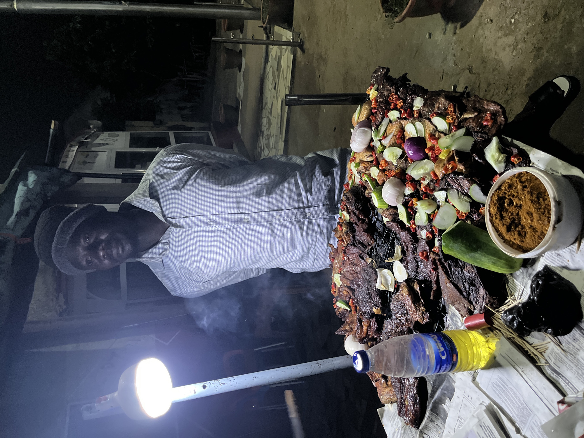 The media file shows a man standing next to a large platter of grilled meat. The meat is dark brown and appears to be seasoned with spices and garnished with chopped onions, cucumbers, and red peppers. The platter is placed on a table covered with newspapers.
The man is wearing a checkered shirt and a dark beanie. He is looking at the camera with a neutral expression. In the foreground, there is a bottle of water and a bottle of oil. There is also a small white container filled with brown spices. Smoke is rising from the meat, indicating it has just been cooked.
A bright lamp is visible in the lower left corner, illuminating the scene. The background appears to be an outdoor setting at night, possibly a street food stall or a backyard gathering. The overall atmosphere is casual and suggests a communal meal or a food preparation event. The context provided indicates the location is Abuja, Nigeria. There is no visible text in the image.