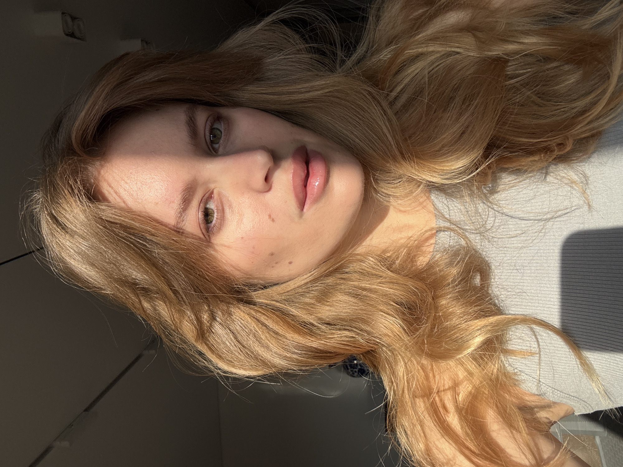 A portrait of a young woman with long, wavy blonde hair, fair skin, and green eyes. The woman is wearing a light gray top. Her lips are slightly parted and appear glossy. Part of her face is illuminated by strong sunlight, while the other side is in shadow. The background is dark. The location, Hrodna, Belarus, is not discernible from the image.