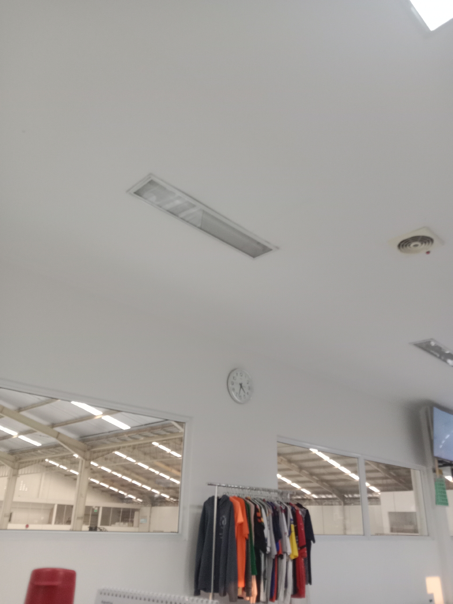 The image shows a ceiling with a fluorescent light fixture and a circular vent. There is a round clock on the wall. Below the clock, there is a rack of clothes. In the background, there is a window looking into a large industrial building with a high ceiling and many lights. The scene appears to be indoors. There are no people visible in the image. The location is not specified as Semarang, Indonesia.