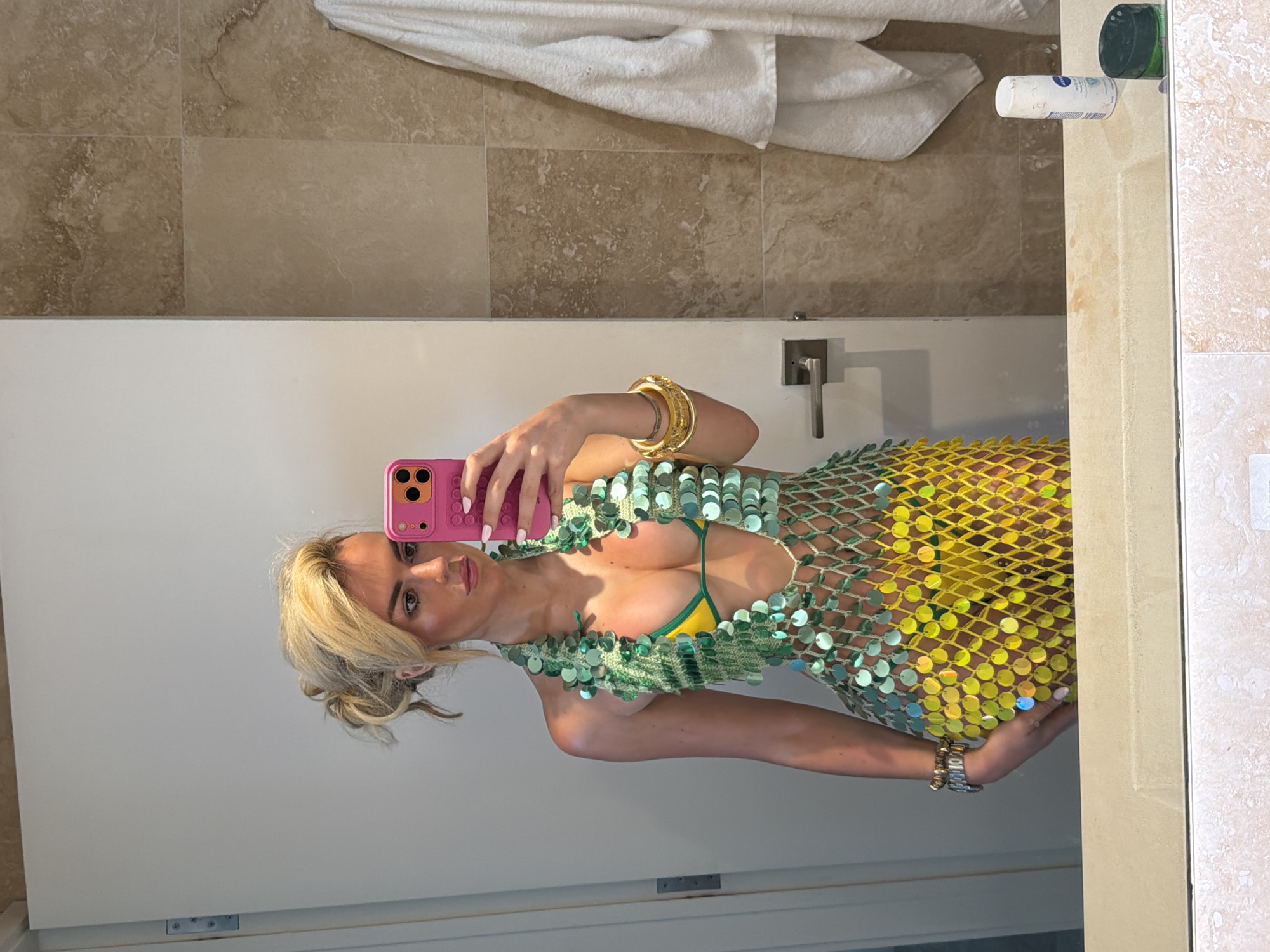 A woman is reflected in a mirror taking a selfie with a pink iPhone. She has blonde hair and is wearing a green sequin top over a yellow bikini top and several gold bracelets. The background appears to be a bathroom with beige marble tiles and a white towel hanging above. A white tube of lotion or cream and a dark green container are visible on a shelf to the right. No specific location is identifiable. Therefore, it is not possible to identify the city of Miami Beach, United States of America, without additional information.