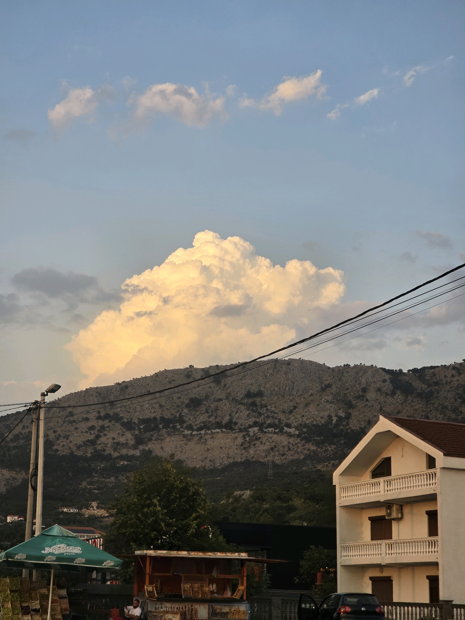 The image captures a scenic view of the city of Podgorica, Montenegro, with a dramatic cloud formation dominating the sky above a mountainous landscape. In the foreground, a white multi-story building with balconies and a dark tiled roof stands to the right. In the lower left, a green umbrella, possibly advertising a local business, shades a stall with various items displayed beneath it. A street lamp is visible on the left side of the frame. A dark-colored car is parked in front of the white building. The background features a rugged, dry mountain slope sparsely covered with vegetation. Several power lines stretch across the upper portion of the image. The overall lighting suggests it is either sunrise or sunset, casting a warm glow on the clouds and the building. A person is seated at the stall under the umbrella, reading.