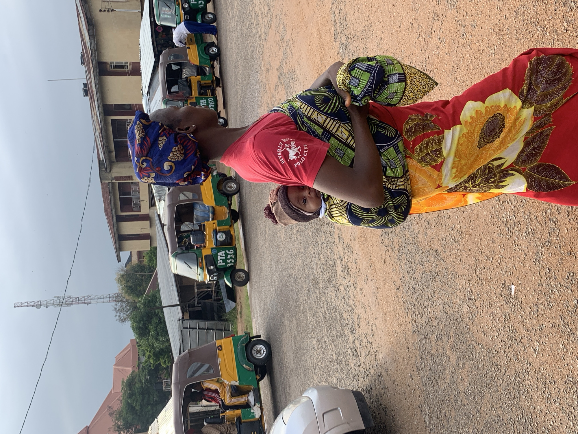 The image captures an outdoor scene in what appears to be a commercial area, possibly a transport hub, in Jos, Nigeria. The main subjects are two individuals, presumably women, and a line of auto-rickshaws, also known as "keke napeps."
One woman is prominently featured in the foreground, facing the camera, though her face is partially obscured by her clothing. She is wearing a vibrant red t-shirt with a logo on the sleeve that reads "BEVERLY HILLS POLO CLUB." She also has a blue headscarf with a patterned design and is adorned with colorful fabric draped over her. The second individual, likely a child, is partially visible behind and to the side of the first woman, looking towards the camera. They are wearing a head covering.
Behind the people, a row of auto-rickshaws are parked. Most are green and yellow, a common color scheme for these vehicles in Nigeria. Some have visible text, including "PTA 1536" on one of the rickshaws. The background reveals buildings and trees, suggesting an urban or semi-urban environment. A tall metal structure, possibly a communication tower, is visible in the distance.
The ground is covered in a gravelly texture. The lighting suggests it is daytime, with bright, overcast conditions. The overall mood is not strongly discernible due to the angle and focus of the image, but the colorful attire of the woman indicates a lively atmosphere. The activity appears to be a pause in transportation services, with the vehicles waiting for passengers.