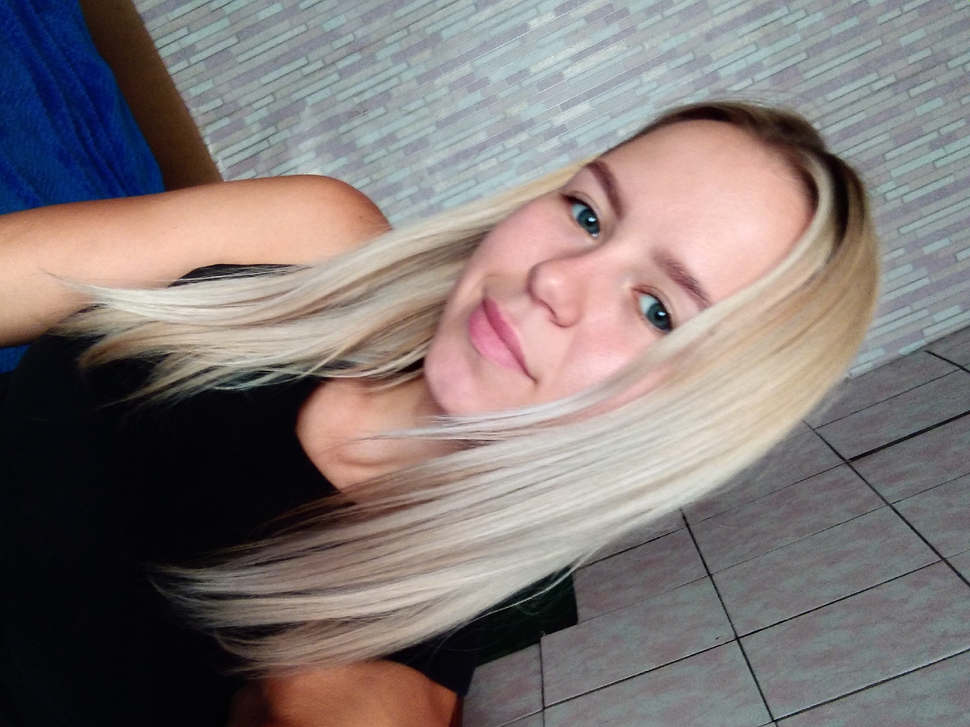 In the city of Radisne, Ukraine, a young Caucasian woman with blonde hair is taking a selfie. She is wearing a black sleeveless top. The background consists of a tiled wall, and to her left, a patterned wallpaper. A blue blanket is visible in the lower left corner of the image.