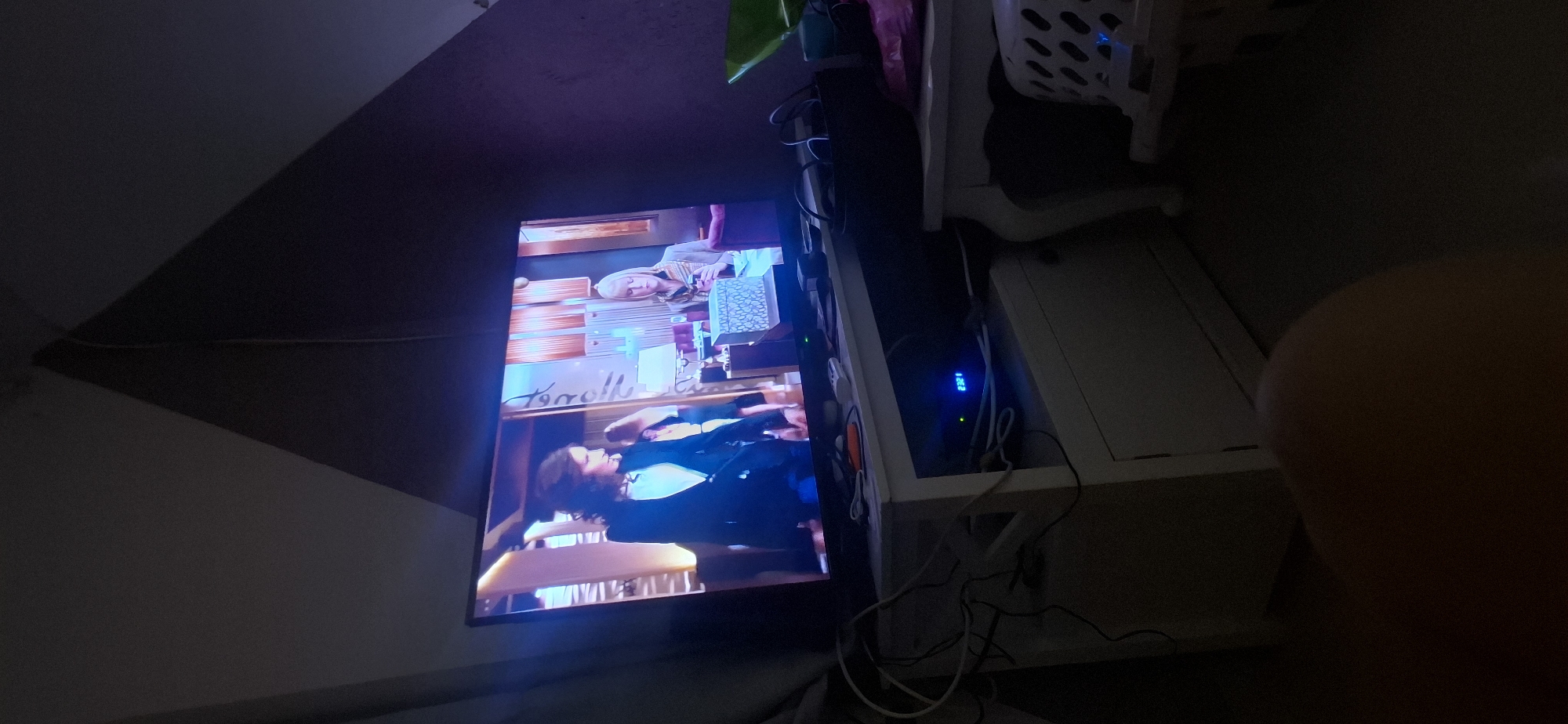 The image shows a television screen displaying a scene with two women. One woman on the left wears a dark blazer over a white top, and the woman on the right wears a hooded garment and holds a phone. The television is placed on top of a white cabinet. Wires are visible near the cabinet, and a digital display on a device below the TV shows "23:21". To the right of the cabinet, a white table with carved legs holds a basket with a white and black patterned insert. The scene appears to be indoors, with a dark, textured wall in the background, suggesting a residential setting in Yerocham, Israel.