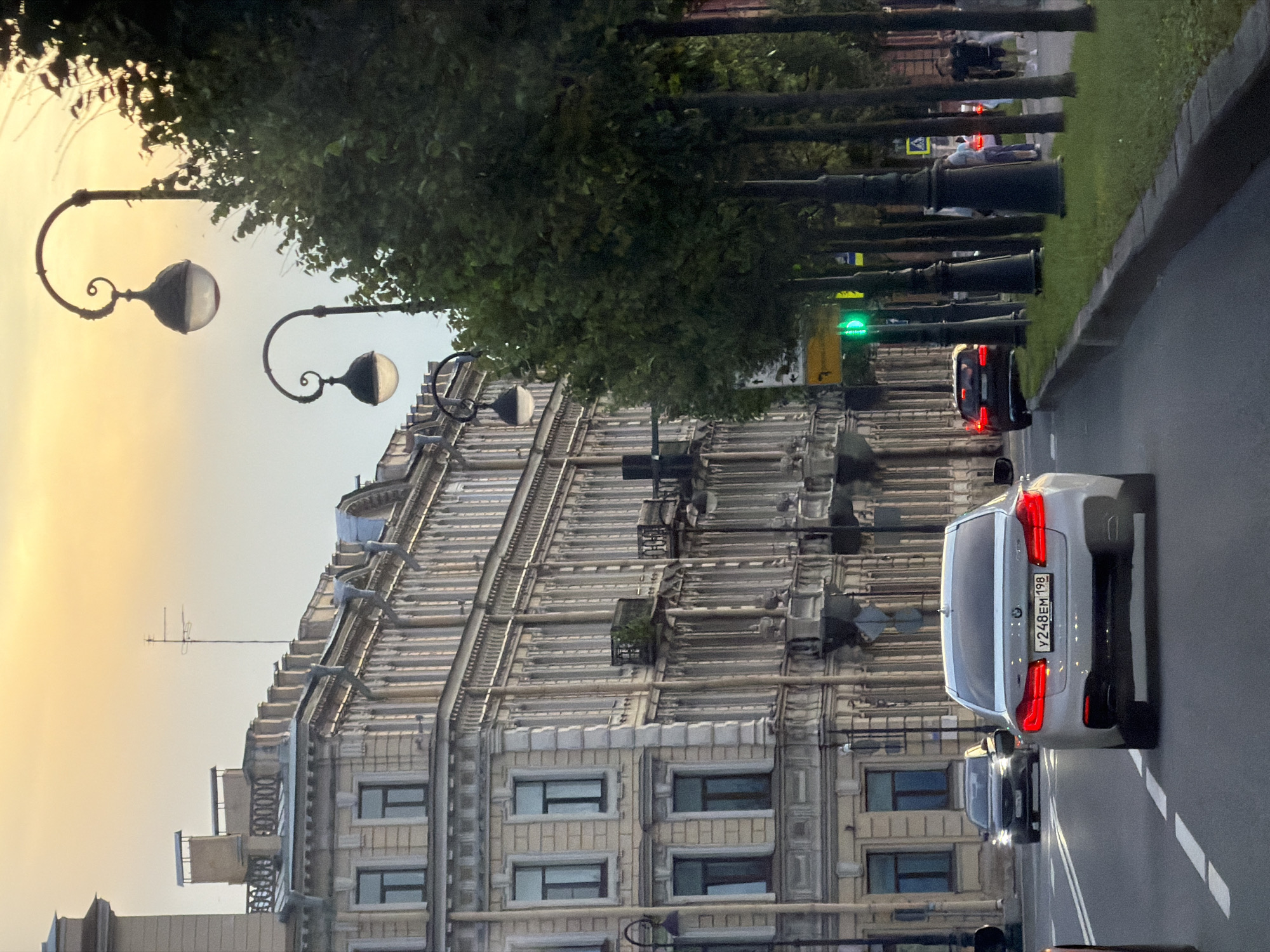 The image captures a street view in Saint Petersburg, Russia, likely during the late afternoon or early evening, as indicated by the warm, low-angled light and the sky's color. The foreground features a gray asphalt road with white lane markings, upon which a silver BMW sedan is driving away from the camera. The car's taillights are illuminated. To the right of the road is a sidewalk with lush green grass and a row of mature trees with dark trunks and dense green foliage. Black lampposts with ornate curved arms and globe-shaped lights line the sidewalk.
Dominating the left side of the frame is a grand, multi-story building with classical architecture. The building is a pale yellow color and features numerous windows, decorative cornices, and balconies. An antenna is visible on the rooftop.
Further down the street, other vehicles are present, including a dark-colored car ahead of the BMW. Traffic signals are visible, with a green light illuminated. Several individuals are seen walking on the sidewalk in the distance, on the right side of the street. The overall impression is a characteristic European city street scene with historic architecture and urban elements.