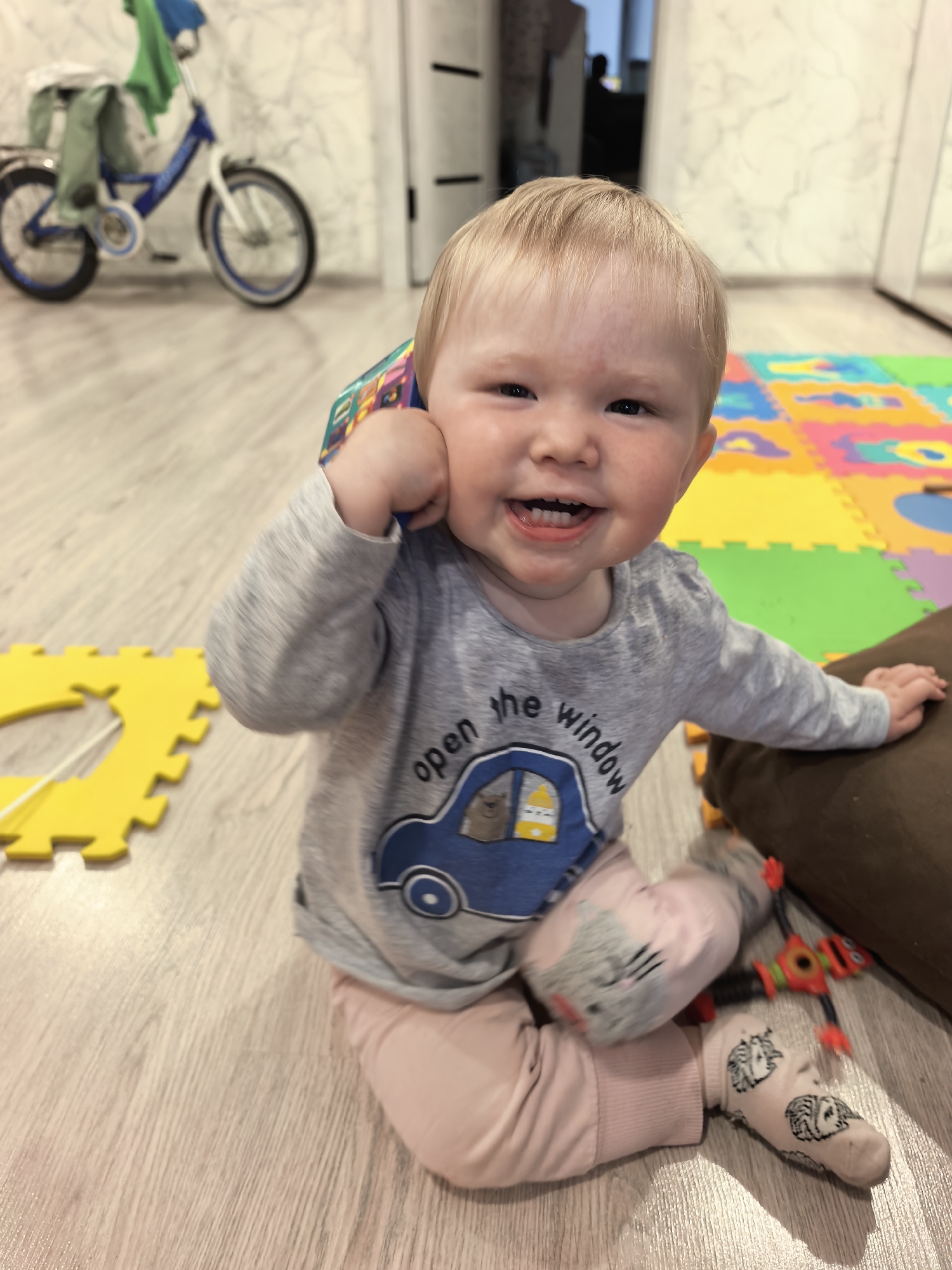 A young Caucasian child with blonde hair and fair skin is sitting on a light-colored wooden floor, looking at the camera and smiling. The child is wearing a gray long-sleeved shirt with a blue car graphic that says "open the window," pink pants, and socks with a cat pattern. The child is holding a colorful object to their ear, appearing to be talking on it like a phone. In the background, a blue bicycle with green fabric draped over it is visible. There are also colorful foam puzzle mats on the floor, as well as a brown pillow and a small toy robot. The environment appears to be an indoor living space, possibly a playroom or living room. There is no indication of the child's location in the city of Sterlitamak, Russia.
