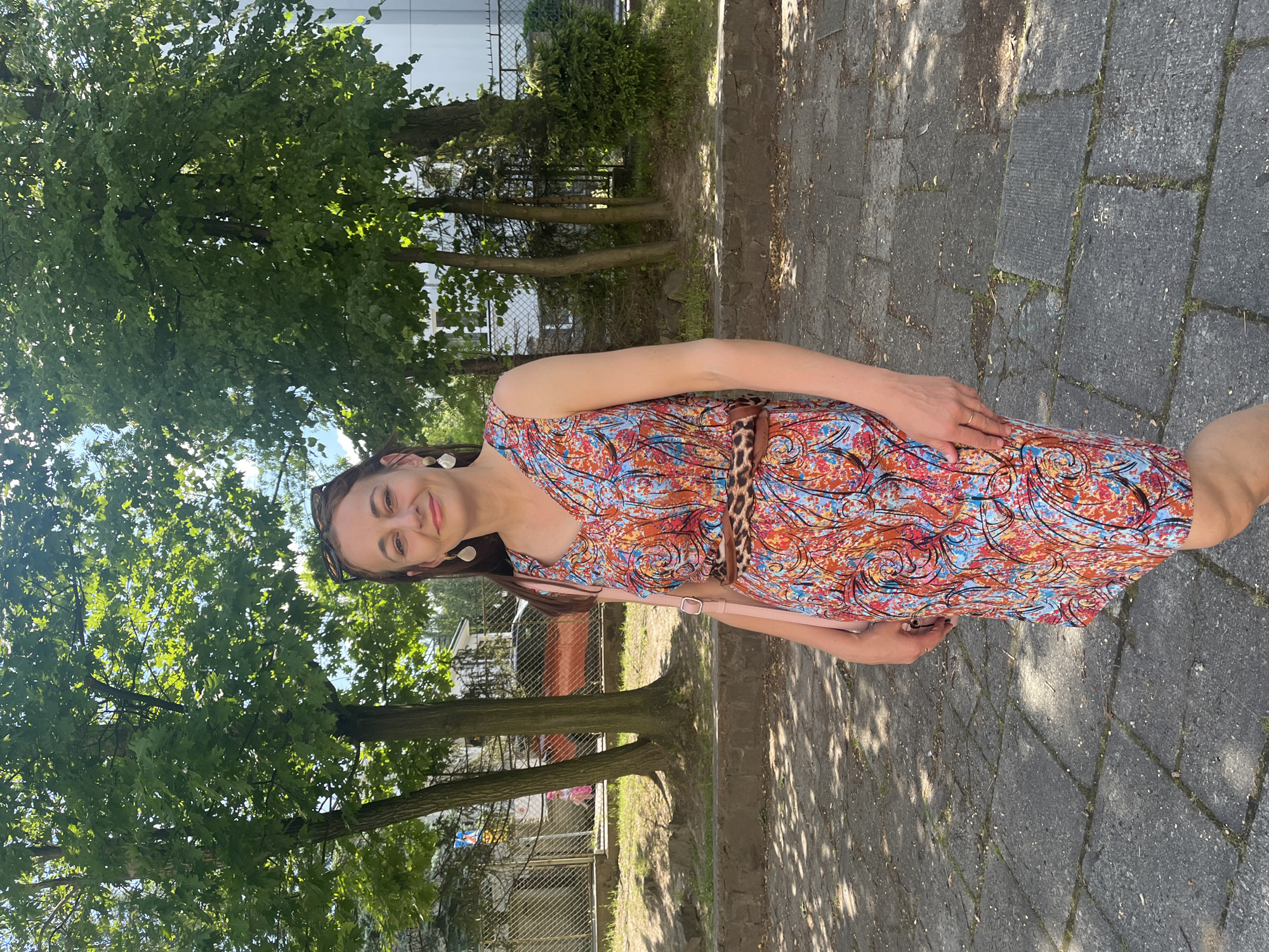 This is a vertical, eye-level, outdoor photograph of a woman standing on a paved walkway. She is smiling and looking towards the camera. She is wearing a colorful, patterned dress with shades of orange, blue, and black. The dress has a V-neck and is cinched at the waist with a leopard print belt. She is also wearing large, white earrings and sunglasses perched on her head. A light pink purse is slung over her shoulder, and her left hand rests on her hip.
The background of the image features lush green trees with dappled sunlight filtering through their leaves. Beyond the trees, there are glimpses of buildings and a chain-link fence. The paved walkway appears to be made of dark gray stones. The lighting suggests it is a sunny day, likely in the late morning or afternoon. The overall mood of the photograph is cheerful and relaxed. There is no visible text in the image.