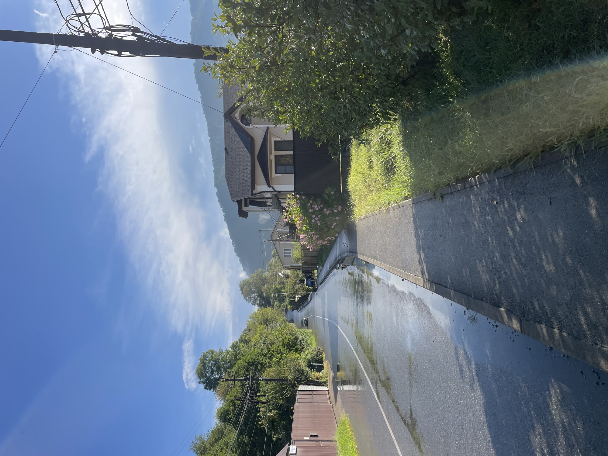The image shows a street scene in Baranovka, Russia, on a bright, partly cloudy day, likely following recent rain. The road is wet and reflects the sky, with a noticeable puddle or stream running alongside the sidewalk on the right. A gray asphalt sidewalk runs parallel to the road. On the right side of the sidewalk, lush green grass and bushes are visible, with a visible rainbow effect cast on the grass. Behind the grass, a modern two-story house with a dark roof and light-colored walls stands. The house has several windows and an arched feature above the upper-level windows. In the background, rolling green hills are visible, partially obscured by trees. A utility pole with numerous wires stands prominently on the right side of the image, extending into the sky. Further down the road, two cars are visible, one dark and one lighter colored, driving uphill. Trees line both sides of the road, and a tall brown fence is seen on the left side of the image, bordering a property. The sky is a vibrant blue with streaks of white clouds.