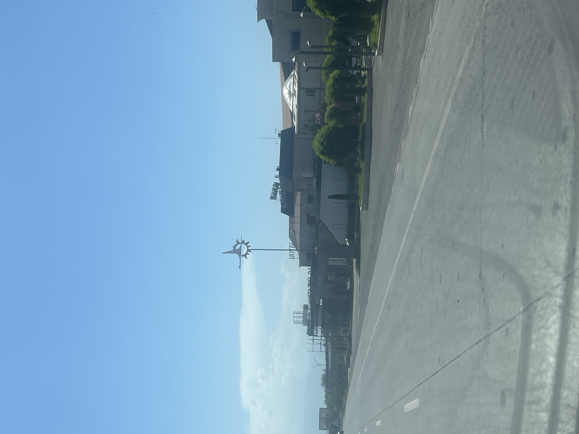 The image depicts a sunlit street scene in Yerevan, Armenia, viewed from a moving vehicle. The bright blue sky dominates the upper portion, mostly clear with a faint wisp of cloud on the left horizon. A wide, grey asphalt road occupies the right side of the frame, featuring a distinct solid white line and faint tire marks.
Along the left side of the road, several multi-story buildings are visible, interspersed with green bushes and small trees. These buildings exhibit varying architectural styles; one prominent structure has a distinctive, light-colored dome roof. Another building, appearing to be commercial, clearly displays the text "HOTEL" on its facade in large, dark letters. Above "HOTEL," partially obscured text that might read "BURGUM" or "BURKAM" is visible. Other structures include streetlights and what appears to be scaffolding or construction elements near some of the buildings.
A striking feature in the mid-ground is a tall pole topped with an intricate, star-like or sunburst ornament, suggesting it could be a decorative streetlight or a small monument. The scene is illuminated by bright daylight, indicating it was captured during morning or afternoon hours, with clear weather. No people are visible, and the overall impression is one of a quiet urban thoroughfare.