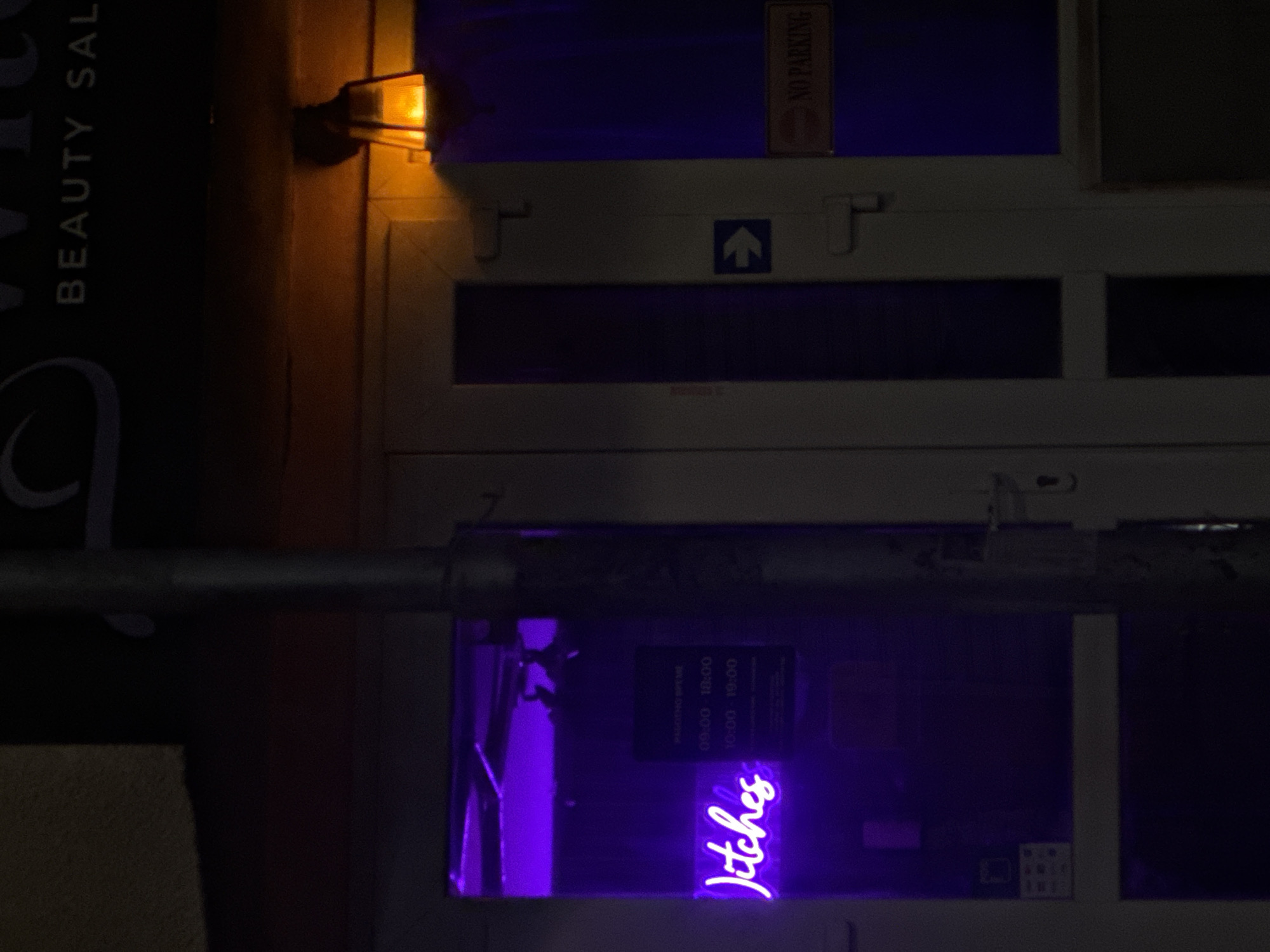 The image presents a dimly lit street scene at night, possibly in Varna, Bulgaria. In the upper left corner is part of a vertical sign reading "BEAUTY SAL,". An old-fashioned-looking lantern glows on the facade of a building. To the right, there's a white frame containing multiple square panes of glass. This frame includes a "NO PARKING" sign and an arrow indicating direction. Inside one of these panes, a purple neon sign reads "Jitches" in cursive. There's also a sign, perhaps a schedule, written in a smaller font. A horizontal metal rod crosses the middle of the frame, obscuring parts of the view.