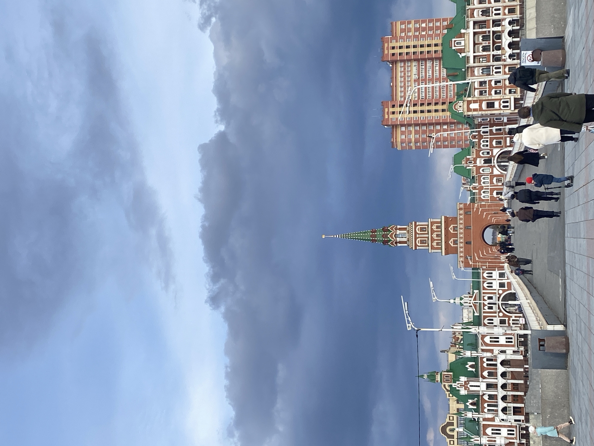 This vertical image captures a vibrant street scene in Yoshkar-Ola, Russia, characterized by its distinctive architecture under an overcast sky. The focal point is a collection of buildings with red-brick facades, white trim, and prominent green roofs, some featuring arched windows and decorative elements. A striking central building boasts a tall, ornate spired tower, reminiscent of traditional Russian or European cathedral styles, with green, red, and white patterns on its spire. In the background, taller, more contemporary-looking apartment blocks with beige or light orange exteriors are visible.
A paved pedestrian area, likely a walkway or square, occupies the foreground and midground. Several people are seen walking along this path, dressed in casual clothing and coats, suggesting cool weather. Their faces are not clearly discernible, but they appear to be going about their daily routines. Objects in the scene include several streetlights and a brown public trash bin located on the right side of the path.
The sky above is predominantly overcast, with various shades of blue-grey clouds indicating a cloudy daytime. The unique architectural style serves as a strong location cue for Yoshkar-Ola, known for its eclectic blend of historical and European-inspired buildings. There is some faint, unreadable text on the trash bin, possibly starting with "M" or "SM". The overall atmosphere is calm and ordinary, capturing a typical urban moment.