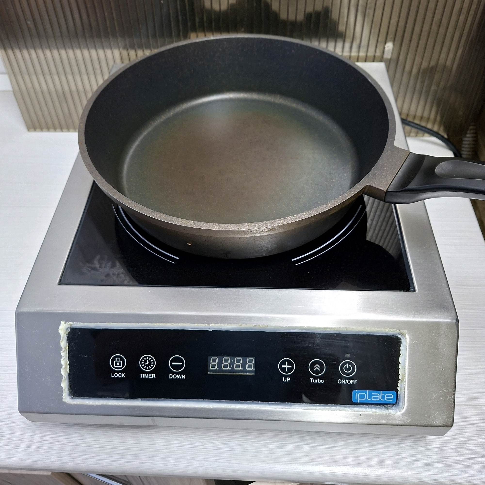 The image shows an induction cooktop with a frying pan on it. The cooktop is a stainless steel unit with a black glass surface. The control panel has icons for "LOCK", "TIMER", "DOWN", "UP", "Turbo", and "ON/OFF", as well as a digital display showing "8.8.8.8". The brand name "iplate" is visible on a blue rectangular label. The frying pan is dark gray and appears to be non-stick. The background consists of a ribbed, metallic surface. The scene is indoors, on a white surface that appears to be a countertop or table. There are no people or animals visible in the image. The location in Anapskaya, Russia, is not discernible from the visual information provided in the image.