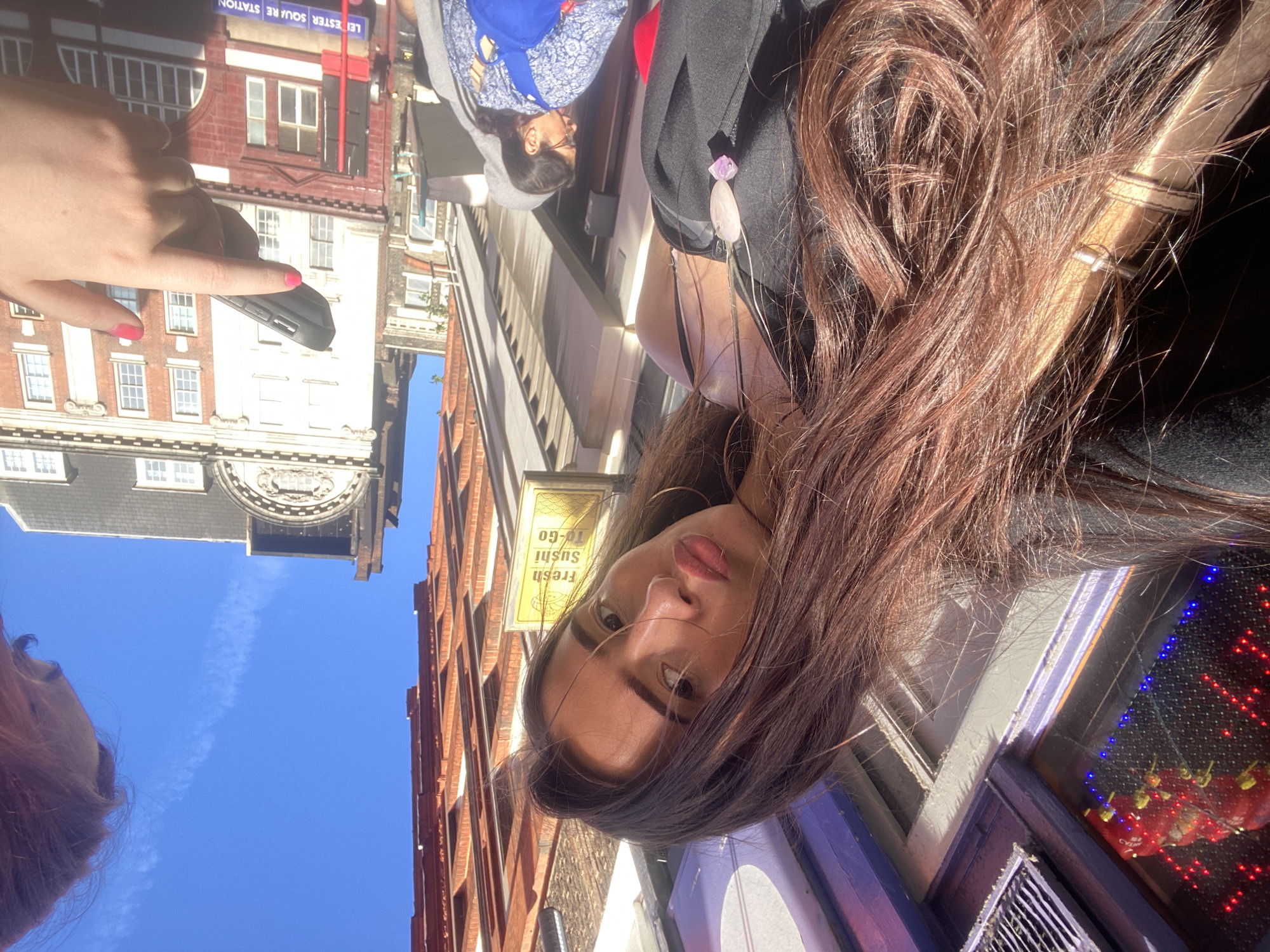 A woman with brown hair is taking a selfie in London. She is standing near a shop with a sign that reads "Fresh Sushi To-Go." In the background, there are buildings with red brick facades and a blue sky above. A hand holding a smartphone is visible, pointing at a sign for "Leicester Square Station." Other people are also visible in the background, but they are out of focus. The overall scene is sunny and vibrant.