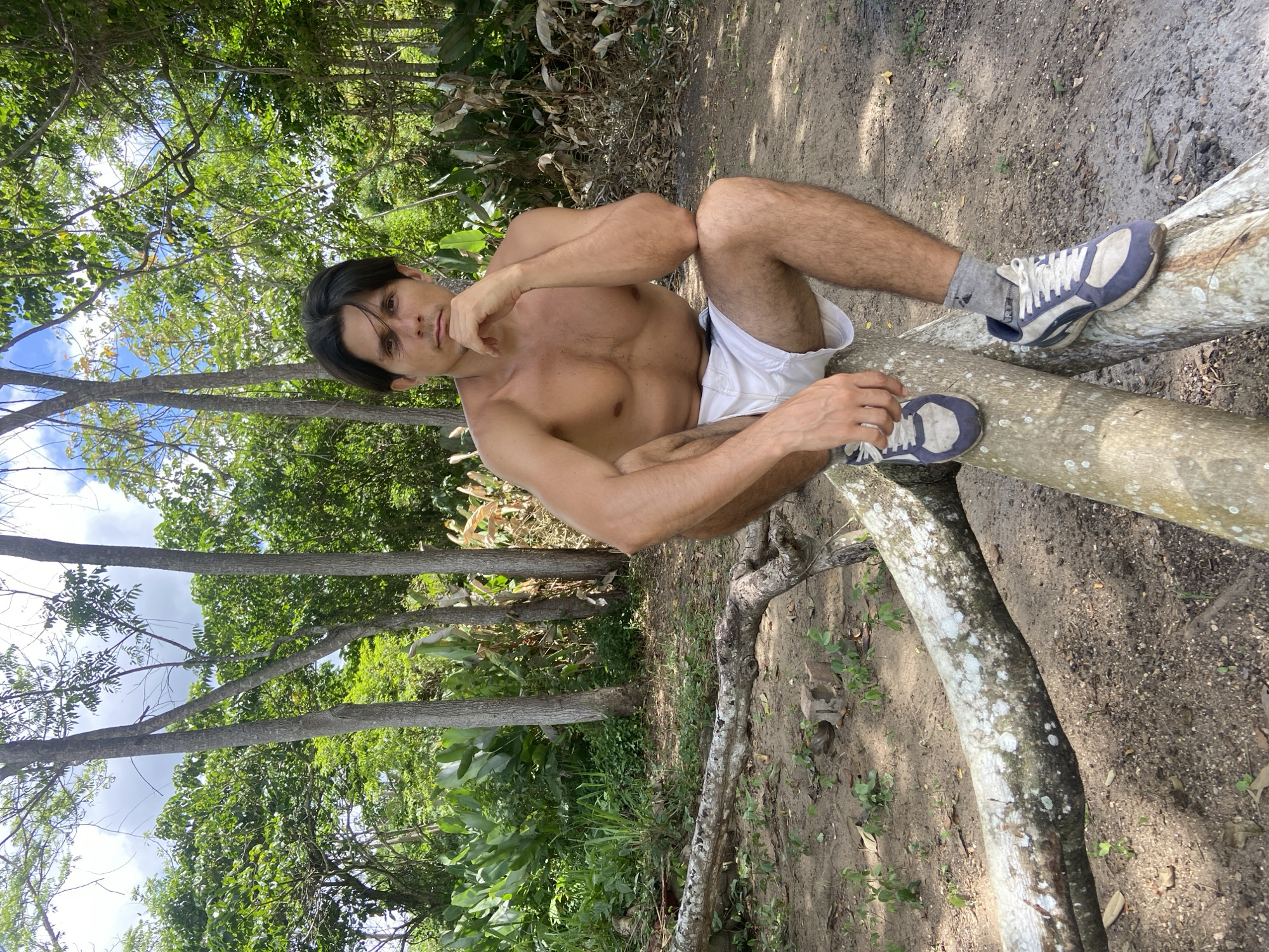 A man is sitting on a fallen log in a wooded area. He is shirtless and wearing white shorts and sneakers. The man has dark hair and is looking at the camera with his hand on his chin. The background is filled with trees and foliage. The ground is covered in dirt and leaves. The scene is set in Los Velásquez, Venezuela.