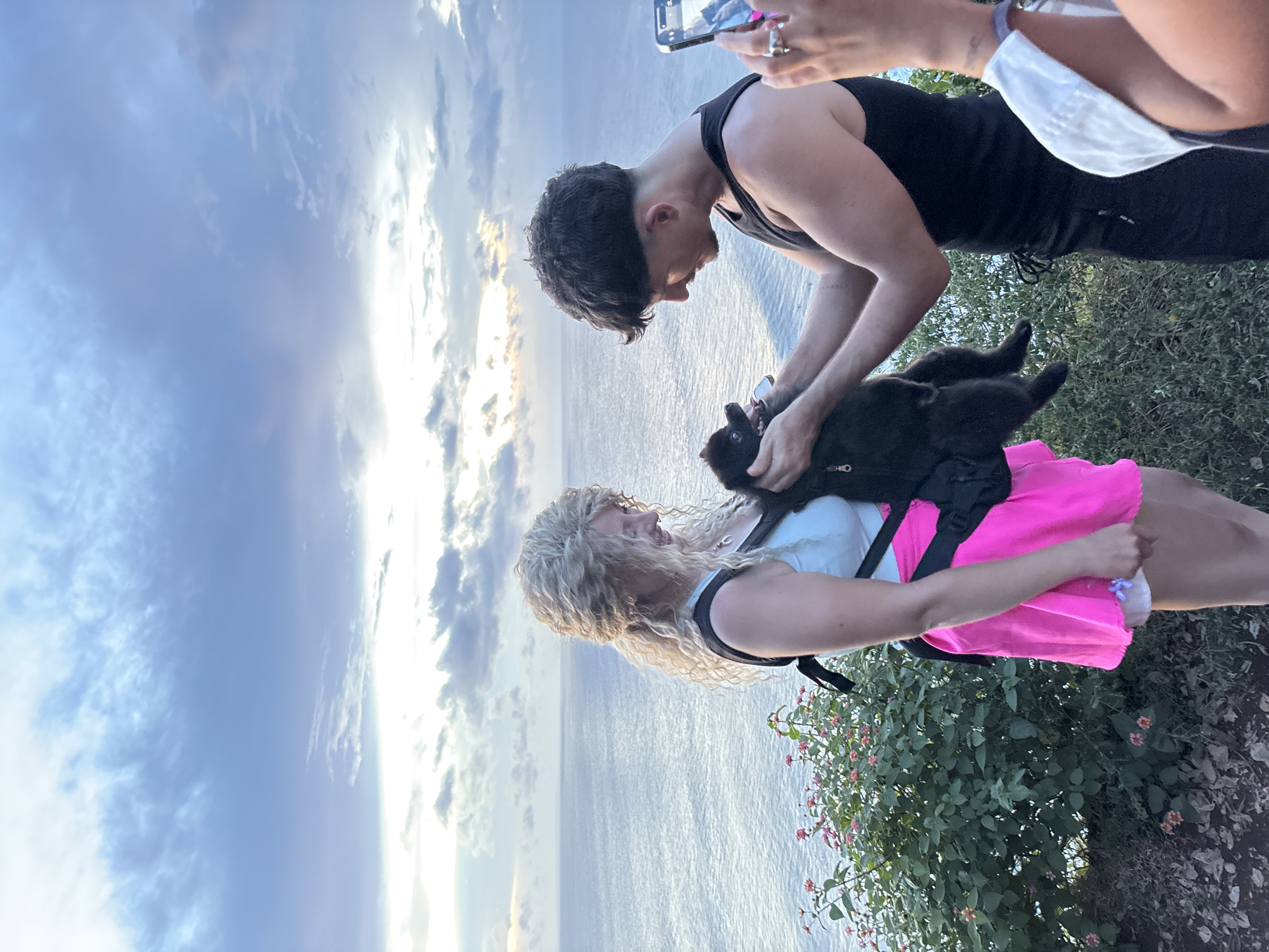 The image depicts a man in a black tank top holding a black puppy above a woman wearing a white shirt, pink skirt, and black backpack. They are standing near a cliff edge, with the ocean visible in the background. A person is photographing the scene with a cell phone. The location is near Uluwatu, Indonesia, evidenced by the landscape and sky. The environment includes green foliage with red and orange flowers. The sky is partially cloudy with a sunset glow.