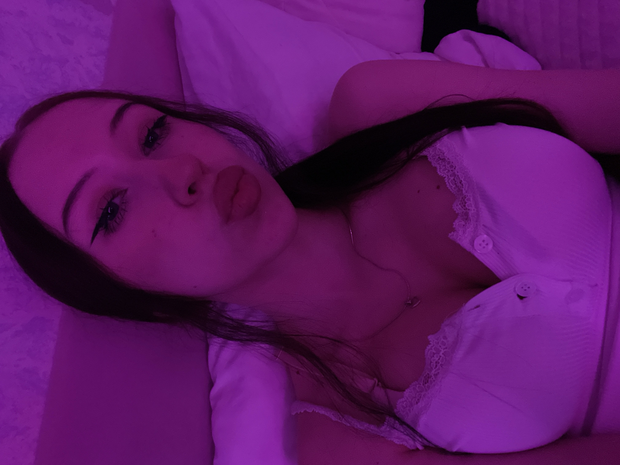 The image shows a close-up, dimly lit selfie of a young woman lying down. She has dark hair, winged eyeliner, and her lips are pursed. She is wearing a white top with lace trim and buttons. The lighting is predominantly pink or purple, casting a shadow over much of the scene. The background appears to be a bed with pillows. There is no discernible information about the location within Votkinsk, Russia.