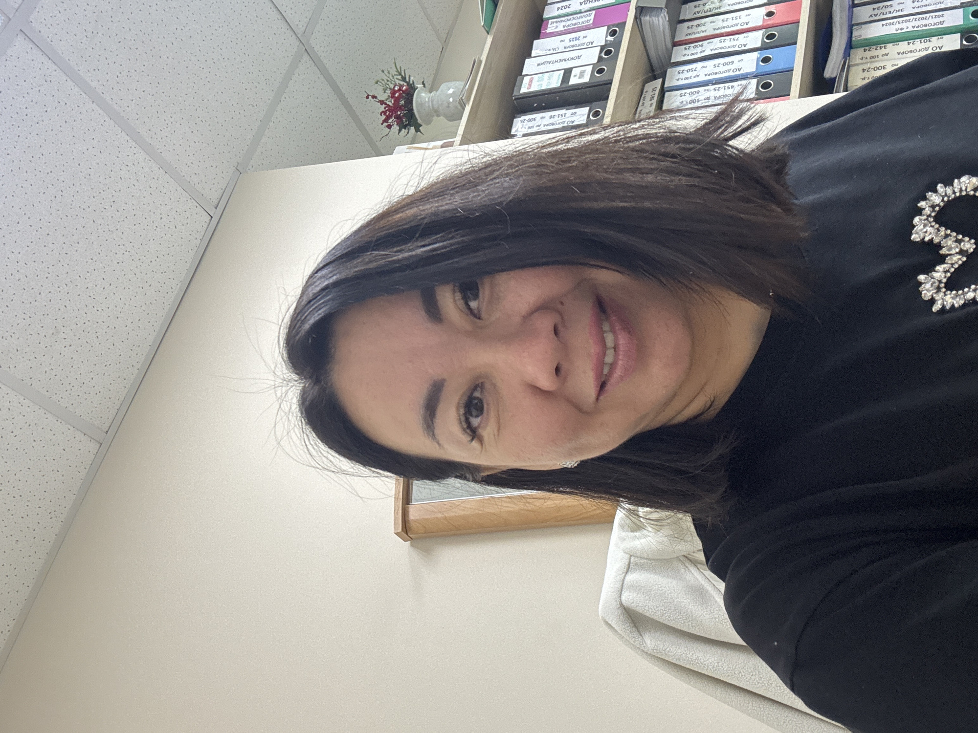 The image shows a medium shot of a woman in an office setting. She has dark brown hair, parted in the middle and falling to her shoulders. She is wearing a black t-shirt with a sparkly embellishment on the chest. She is smiling and looking directly at the camera. In the background, there is a wall, a ceiling with acoustic tiles, a shelf filled with binders, and a small decorative plant in a vase. To the left of the woman, a light-colored jacket or coat is draped over a chair. The overall impression is that of a casual selfie taken in an office. The image does not provide any specific details about the location within the city of Yakty-Kul, Russia.