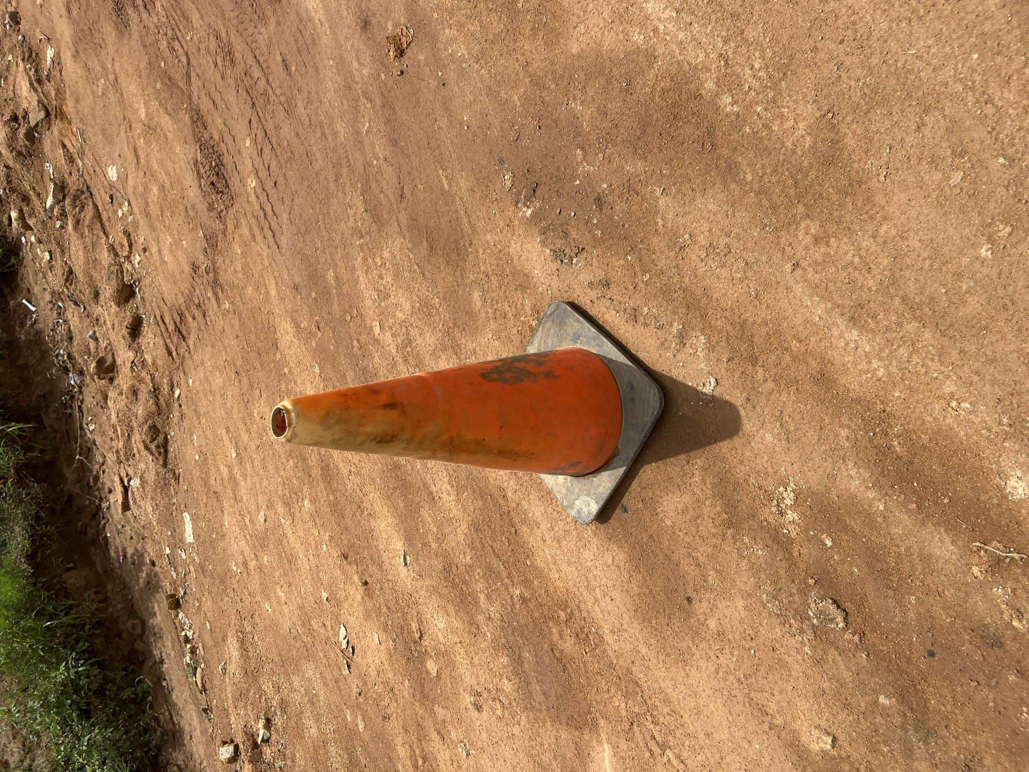 An orange traffic cone with a black square base sits on a dirt road. The road surface is reddish-brown and appears to be compacted dirt with some loose pebbles and debris. Tire tracks are visible on the road surface. In the background, there is a grassy embankment with some scattered litter. The scene is likely on a road in Bukuru, Nigeria, as suggested by the color of the soil, vegetation, and tire track patterns common in the region.