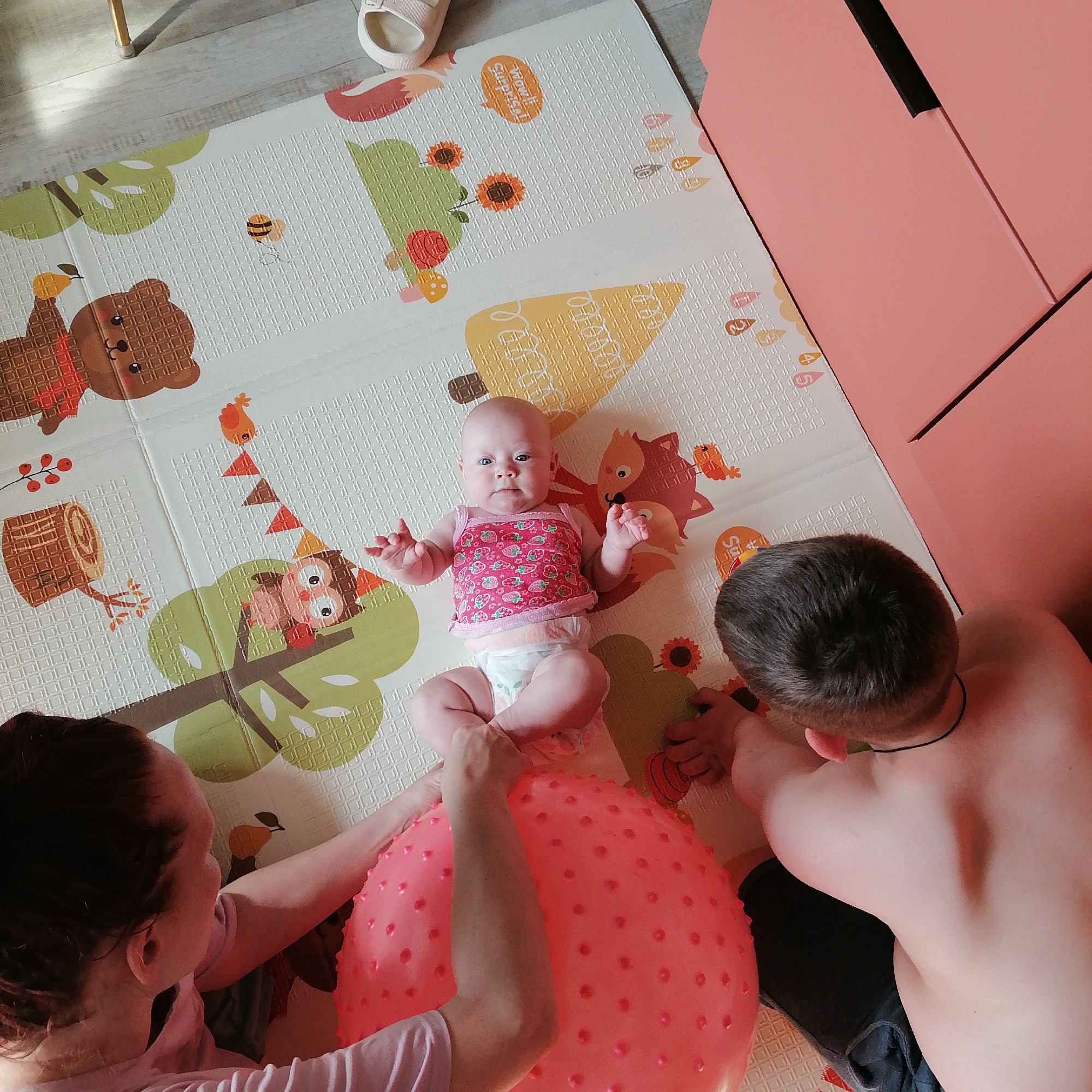 In Trubacheyevka, Russia, on a soft, printed mat designed with cartoon animals and nature motifs, a family is engaged in playtime. A baby, wearing a pink strawberry-patterned top and a diaper, lies on its back, looking up with wide blue eyes. Two adults are positioned around the baby. A woman, with reddish-brown hair and wearing a pink t-shirt, is on the left, her arms reaching towards the baby. A man, with dark hair and bare-chested, is on the right, leaning over the baby. A large, spiky pink ball is centrally located on the mat, with the baby's legs resting on it and the adults' hands interacting with it. The background shows a portion of a pale wooden floor and a tall, peach-colored cabinet. The lighting suggests an indoor setting, possibly a living room or playroom.