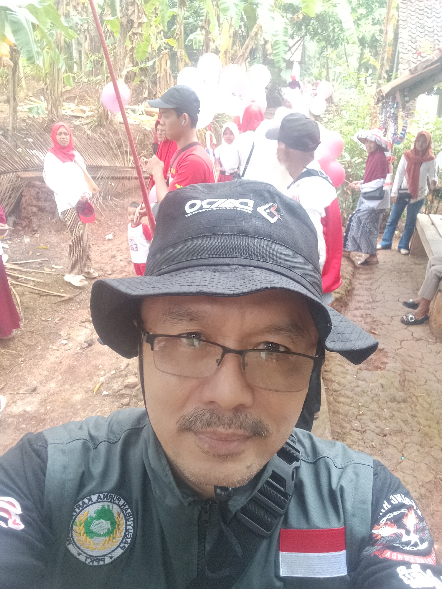 The image is a selfie taken outdoors in Bojongmengger, Indonesia, likely during a local event or gathering. The foreground features a man wearing a black bucket hat, glasses, and a gray jacket with patches and an Indonesian flag. He has a mustache and is looking at the camera. In the background, several people are visible, some wearing traditional Indonesian attire like hijabs. There are also balloons and what appear to be bamboo structures, suggesting a festive or community setting. The ground is dirt and appears somewhat uneven. The overall impression is that of a candid moment captured during a public event in a rural or semi-rural Indonesian location.