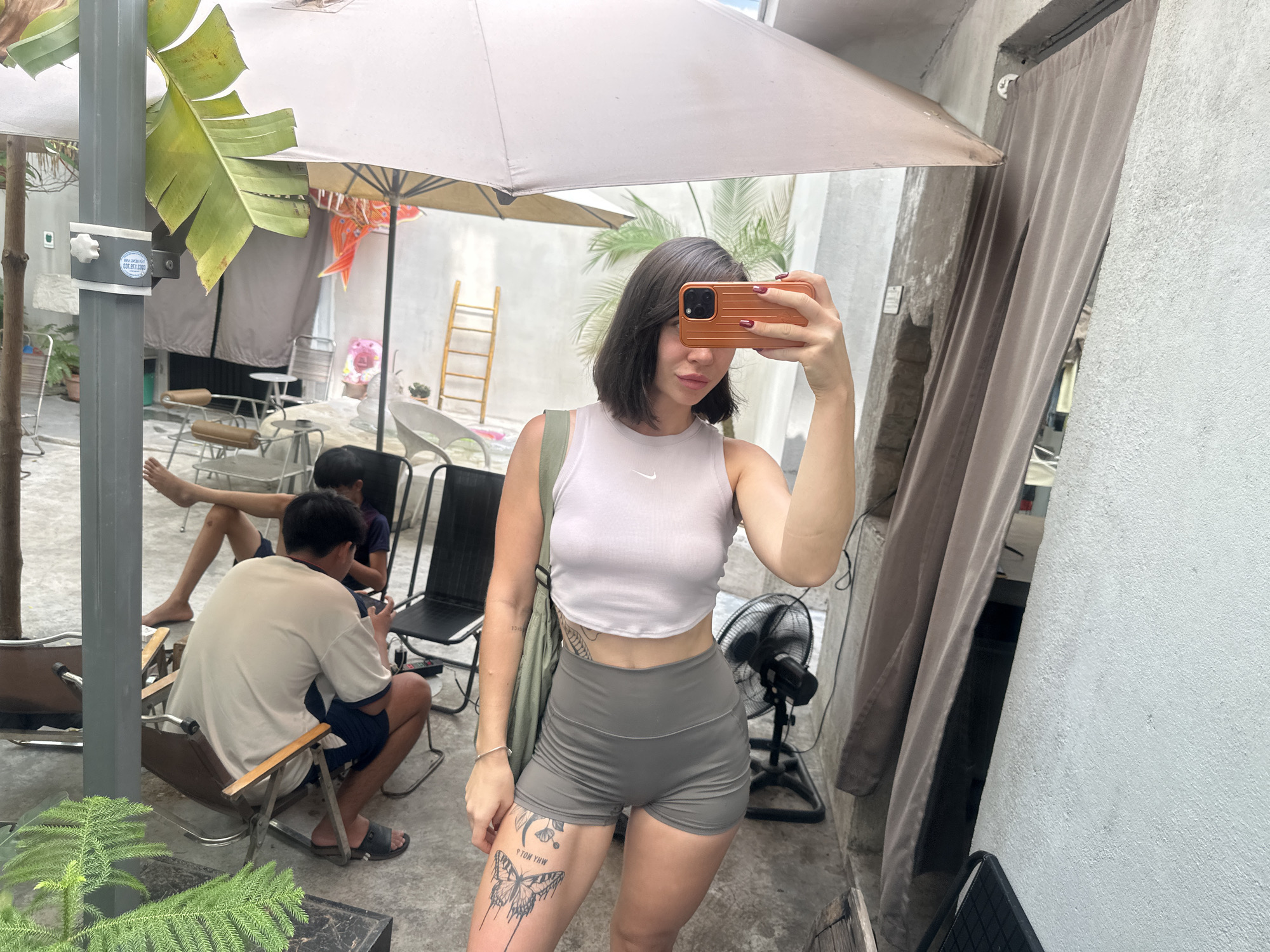 The image depicts an outdoor seating area of a cafe, possibly in Bac Nha Trang Ward, Vietnam, with a woman taking a mirror selfie. She is wearing a light purple crop top and grey shorts, with a large butterfly tattoo on her left thigh. The background shows several patrons seated at outdoor tables, with a fan and a ladder visible. The overall atmosphere appears casual and relaxed.