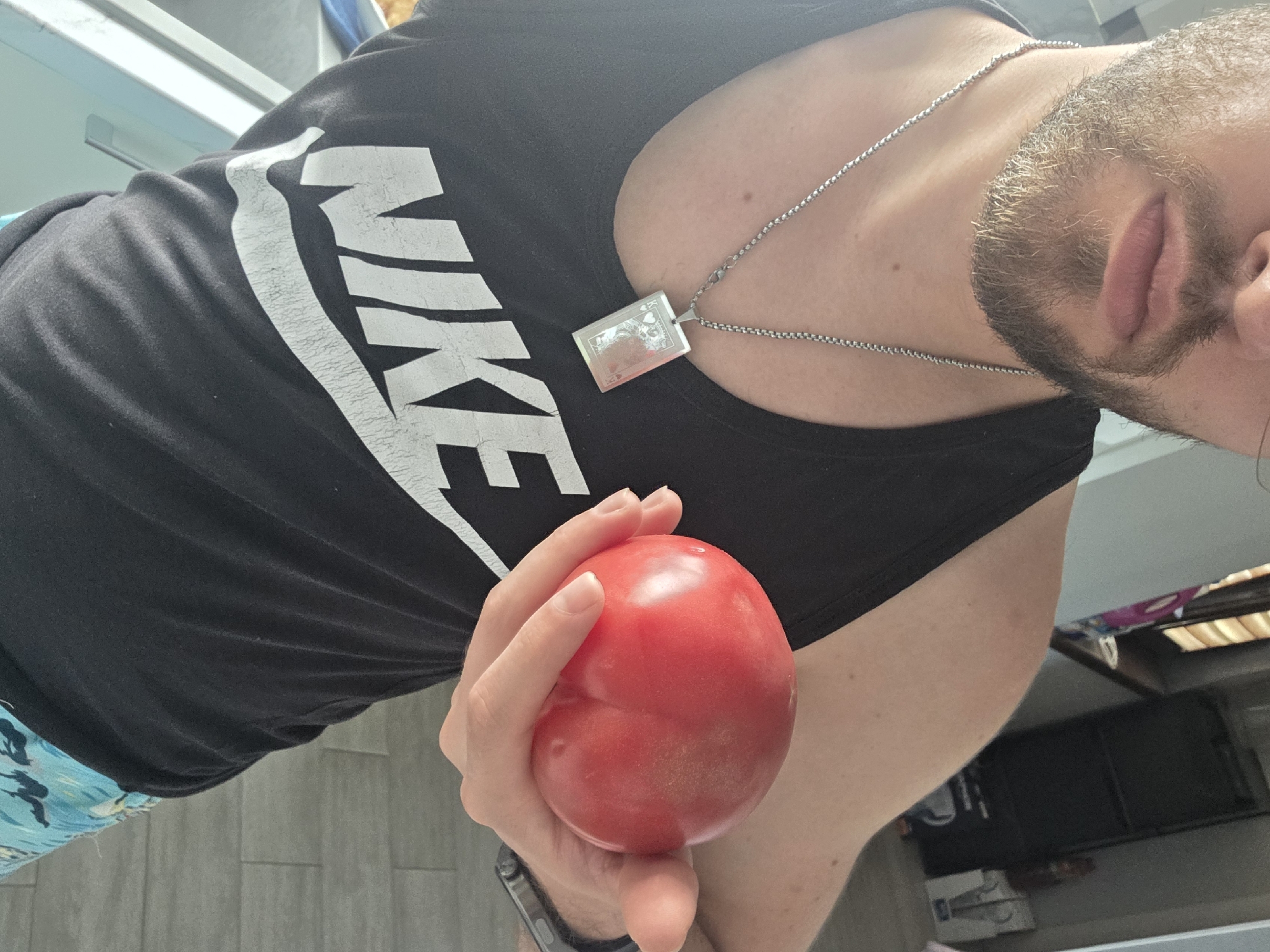 A medium shot shows a person holding a red tomato in their left hand. The person is wearing a black tank top with a white "NIKE" logo on the chest, a silver chain necklace with a rectangular pendant, and an apparent fitness tracker watch on their left wrist. The person has a light beard and visible skin tones. The background is slightly out of focus, revealing a glimpse of an interior space with tiled flooring and some indistinct objects. There is no information about the location in the city of Yagodovo, Bulgaria.
