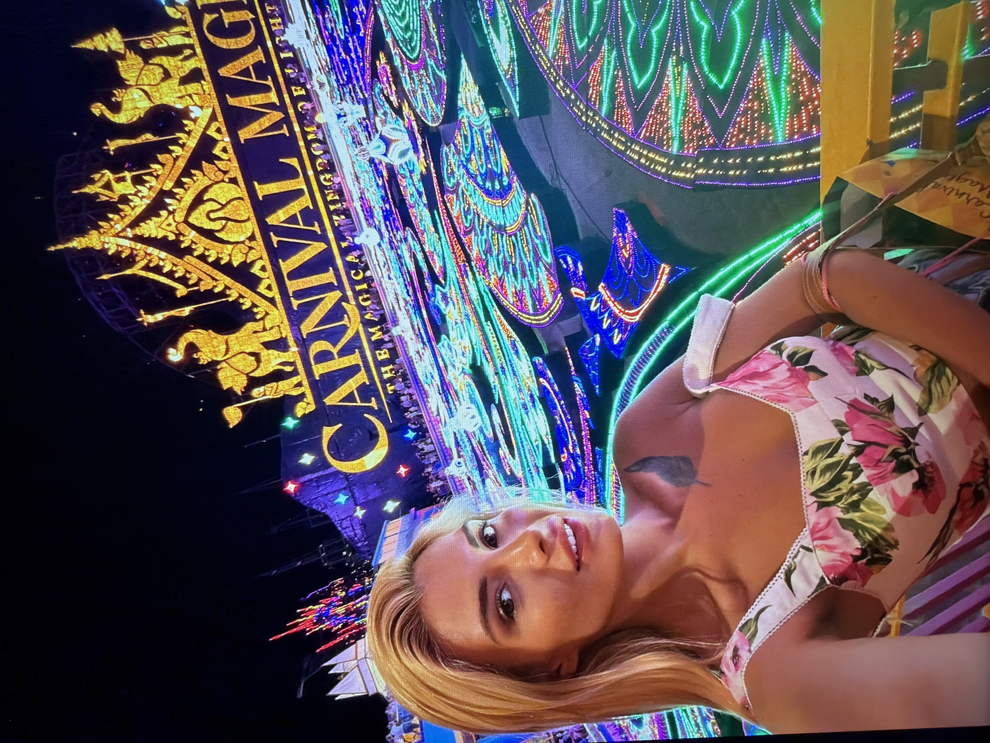 The image features a woman posing for a photo at night, illuminated by a vibrant light display. The primary subject is a woman with blonde hair, wearing a white top with a pink and green floral pattern. She has a tattoo on her left shoulder/upper chest and bracelets on her right wrist. She is looking directly at the camera with a slight smile, suggesting a selfie or close-up portrait.
The background is dominated by an elaborate, colorful light festival or carnival display. To the left, there's a large golden sign or structure featuring intricate patterns and figures, lit brightly against the dark night sky. Beyond this, a sprawling array of LED lights forms diverse geometric and decorative patterns in vivid hues of blue, green, purple, and red, creating a magical atmosphere.
The time of day is clearly night, as indicated by the darkness surrounding the brightly lit installations. The visible text on the golden sign reads "CARNIVAL MAGICAL KINGDOM OF LIGHTS," with "THEM MAGICAL KINGDOM" also partly legible below. The event is taking place in Hamburg, Germany, and appears to be a festive outdoor light show.
