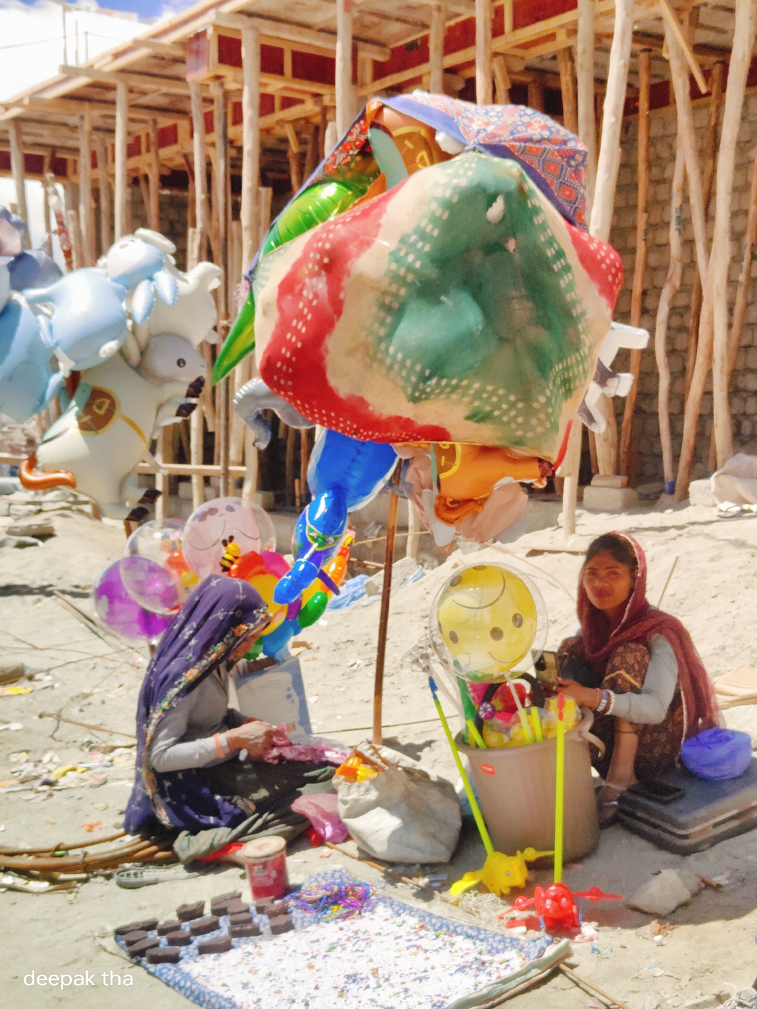The image depicts a street scene in Leh, India, with two women selling balloons and other small items. In the foreground, a woman in a purple headscarf and grey top is sitting on the ground, appearing to be engaged in arranging or selling goods laid out on a patterned cloth. To her right, another woman wearing a maroon headscarf and patterned outfit is also seated, looking towards the camera and holding something in her hands.
Behind the women, a collection of colorful balloons are displayed, including large inflatable animals like a white horse and blue creatures, as well as smaller round balloons with smiley faces and other designs. Some of these balloons are attached to sticks held upright in a bucket. In the background, there is scaffolding and wooden structures, suggesting ongoing construction or market stalls. The ground is sandy and strewn with debris. The overall atmosphere suggests a bustling market or a roadside vendor operation in the arid environment of Leh.