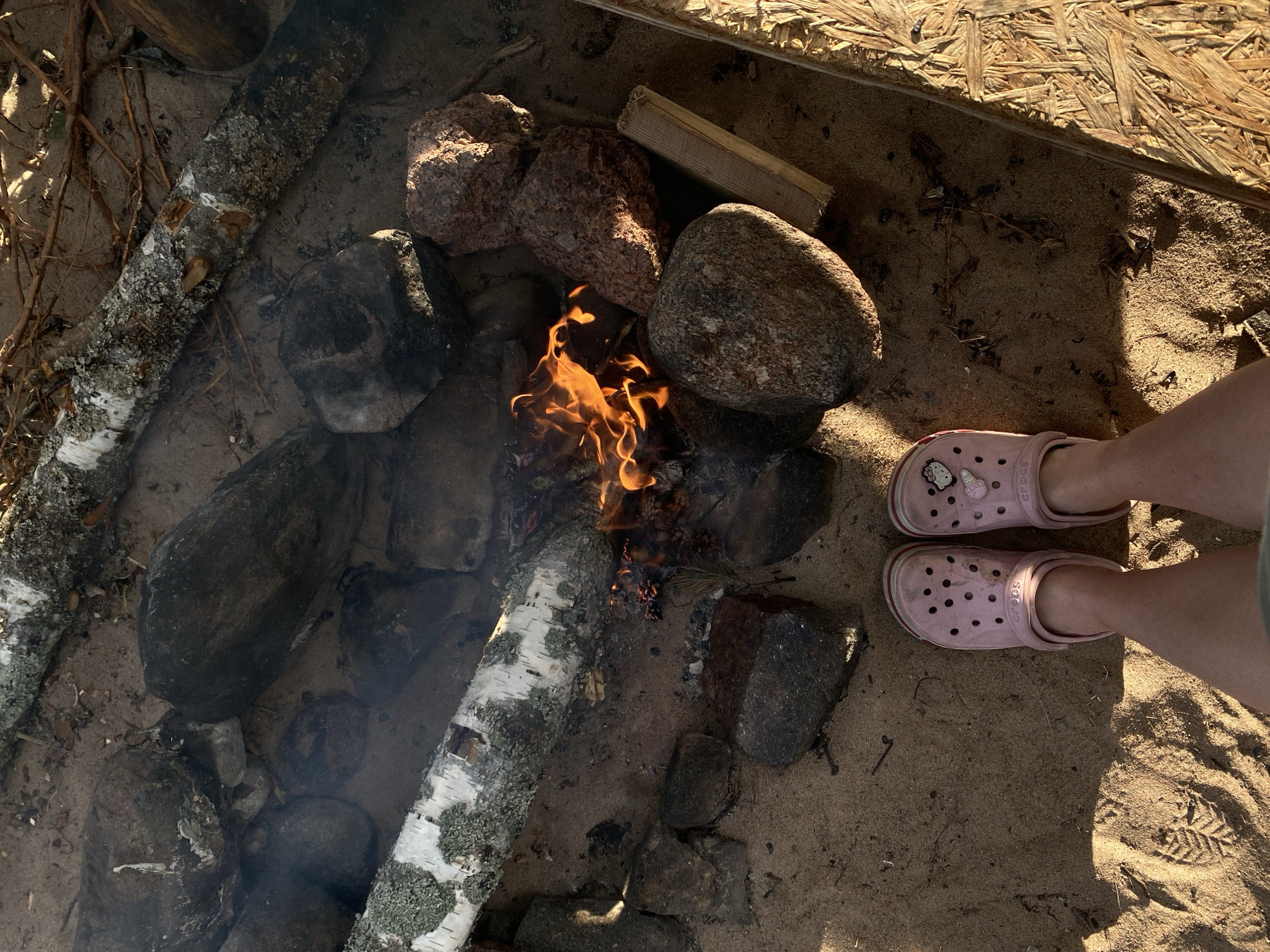 The image depicts a campsite on sandy ground, likely in Baltiiskoe, Russia, as suggested by external context. A small campfire burns in the center, surrounded by several large rocks and pieces of birch wood. A pair of pink Crocs, adorned with a Hello Kitty charm on the left shoe, are positioned on the sand in the foreground, with legs wearing them visible from the knees down. The ground is sandy with scattered debris, including small twigs and pinecones. Sunlight casts strong shadows, indicating it is either morning or late afternoon. The scene suggests a relaxed outdoor setting, possibly a beach or a forest clearing near water.