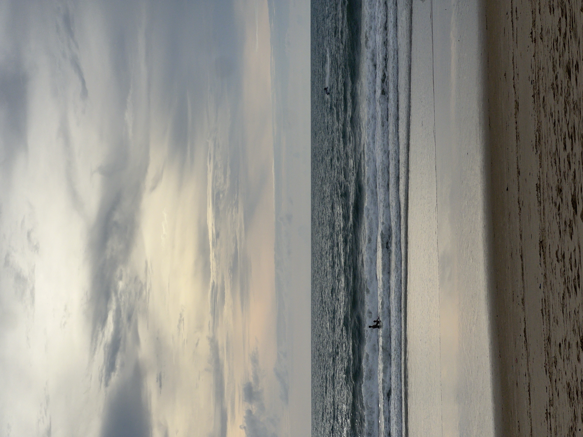 The image captures a serene beach scene at Surin Beach in Choeng Thale, Thailand, under an overcast sky. The ocean is a deep, choppy blue, with white-capped waves rolling towards the shore. In the middle distance, two figures, likely children or adults in shallow water, are silhouetted against the waves. One is standing, and the other appears to be sitting or crouching. Further out in the ocean, a jet ski can be seen leaving a faint wake. The wet sand of the beach reflects the muted colors of the sky and sea. The foreground consists of dry, textured sand with scattered debris.