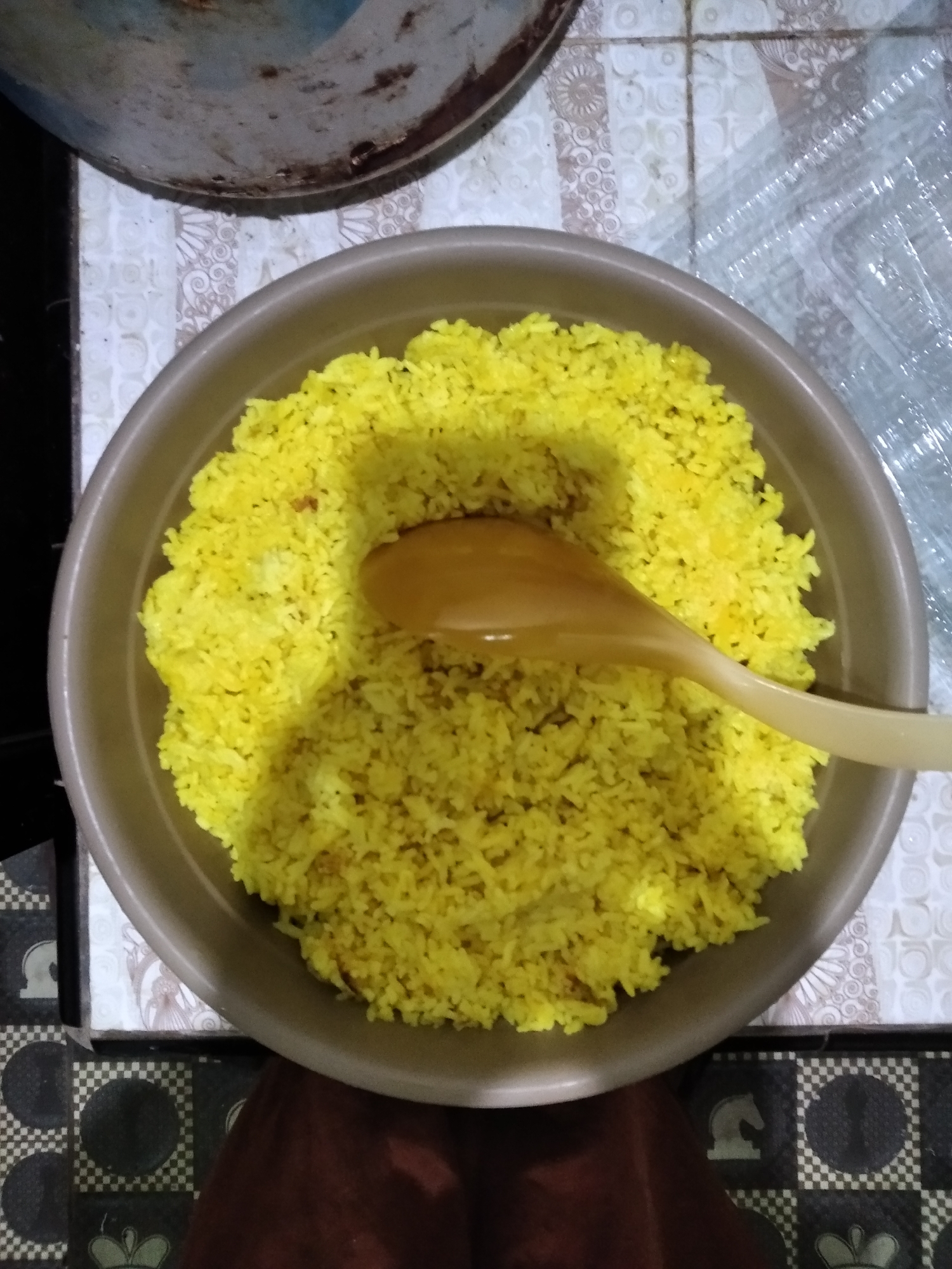 A light brown plastic bowl filled with bright yellow rice is centered in the image. A light brown wooden spoon is placed in the rice, with its handle extending out of the bowl towards the right. The bowl sits on a patterned surface, part of which appears to be a tablecloth with a white and gray floral design, and another part has a black and white checkered pattern. In the upper portion of the image, a portion of a large, metallic pot with some rust is visible. The setting appears to be a kitchen or dining area. The image was taken in Rambipuji, Indonesia.