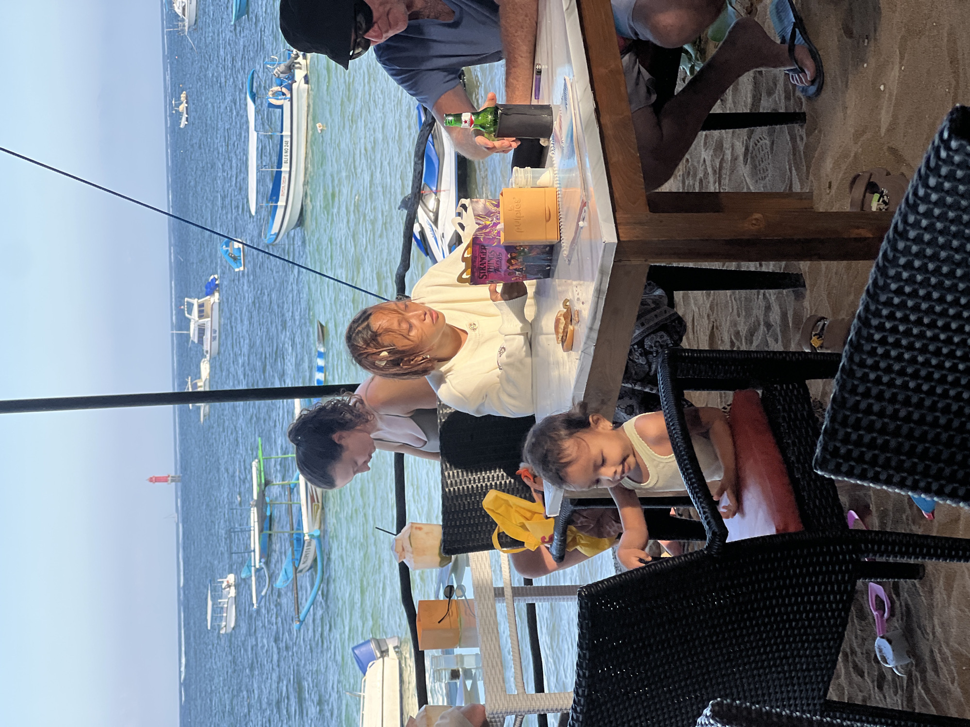 At a beachside establishment in Denpasar, Indonesia, a group of individuals are seated at a table on the sand, with the ocean and boats visible in the background. A young child, wearing a light-colored tank top, is seated in a black wicker chair and appears to be interacting with something on the table. To their right, a young woman with braided hair, dressed in a white long-sleeved shirt, is looking down at a box. A man with a black hat and sunglasses, wearing a blue t-shirt, is seated to the woman's right, with his hand reaching towards a bottle of beer in a black holder. A white table with a marble-like surface is in the foreground, laden with food and drinks, including a half-eaten sandwich and a bottle of beer. A woman with dark curly hair, wearing a dark top, is seated to the left of the young woman, looking away from the camera. The environment is sandy, with black wicker chairs and tables scattered around. Several boats are anchored or moving in the calm, blue water of the ocean under a clear sky.