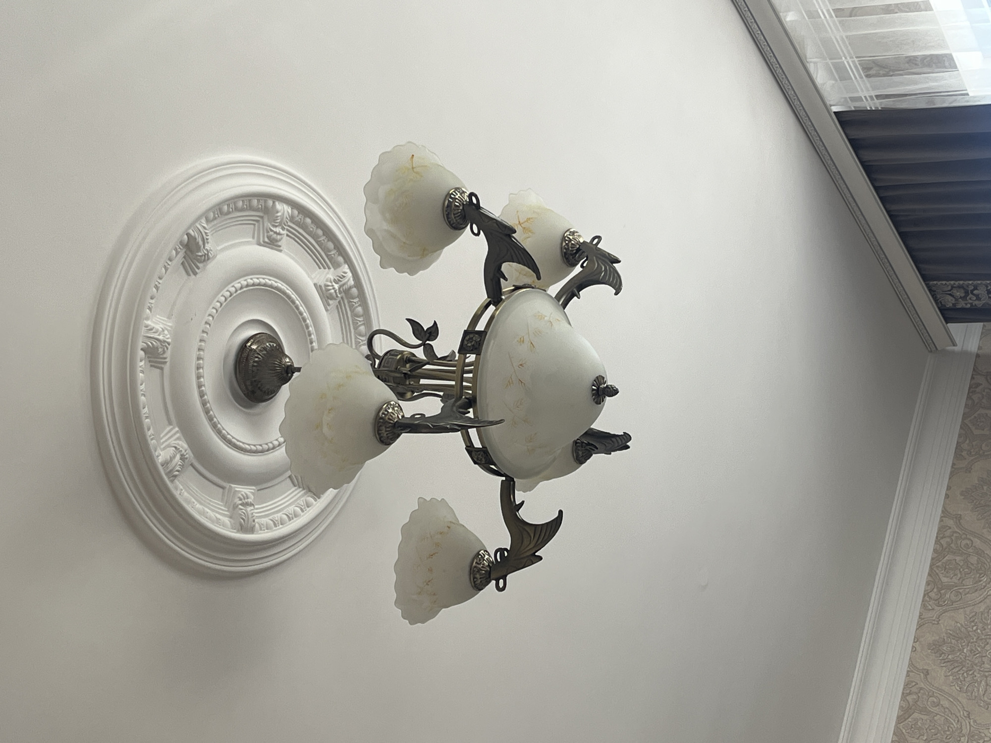 A low-angle shot depicts an ornate, vintage-style chandelier affixed to a circular ceiling medallion in a room in Kaspiysk, Russia. The chandelier has a bronzed metal frame supporting five frosted glass shades with a floral design and scalloped edges. Each shade is mounted on a decorative arm resembling a stylized bird or dragon. The ceiling and wall are painted a bright white. A section of the wall to the right is decorated with patterned wallpaper. In the upper right corner, blinds and an ornate window frame are visible. The setting appears to be an interior space, possibly a residential or commercial building.