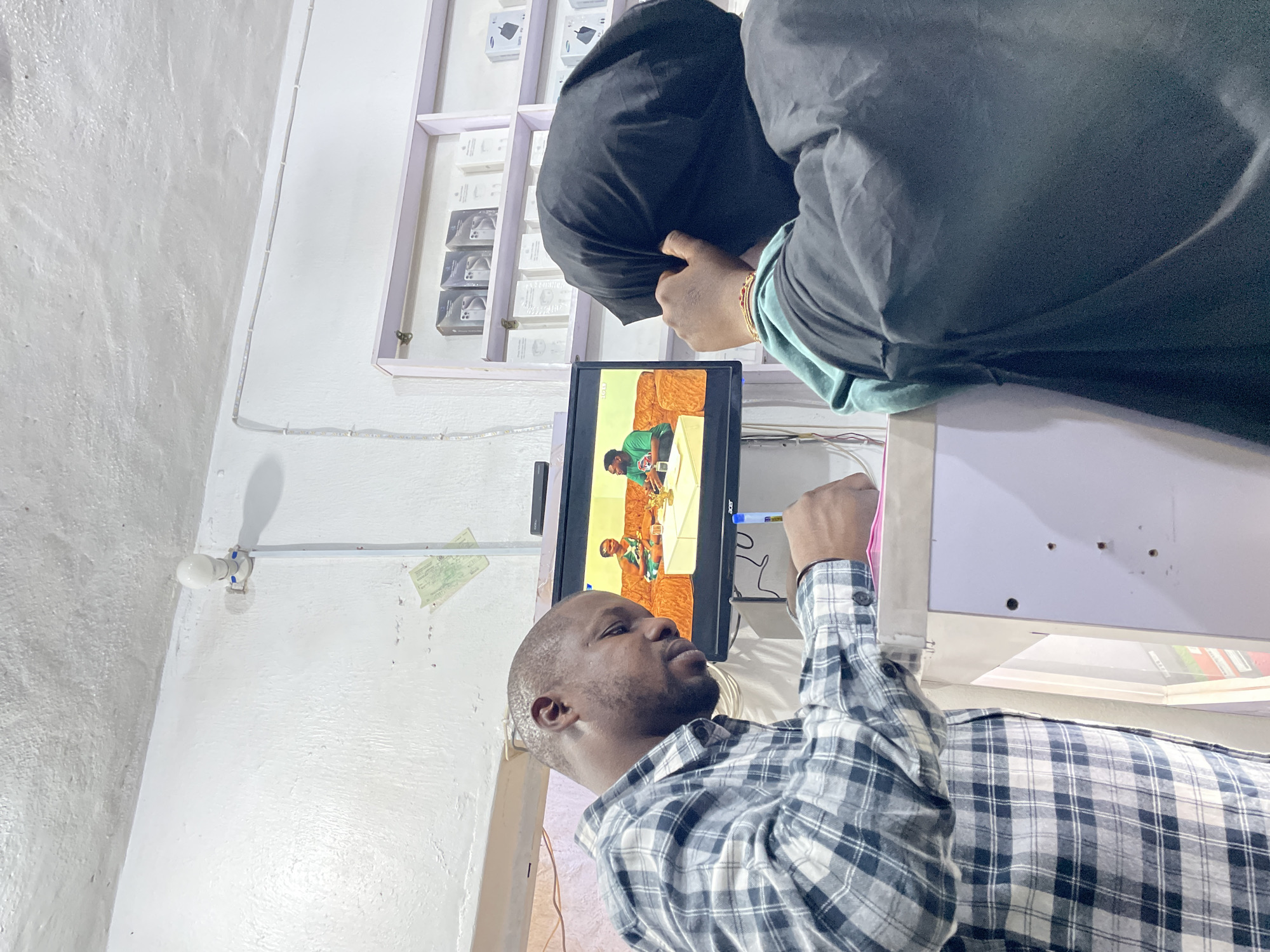 This image captures an indoor scene, likely a small electronics or accessory shop in Jos, Nigeria. In the foreground, a man with a shaved head and a short beard, wearing a blue and white plaid shirt, looks intently upwards at a small flat-screen monitor. His right arm is raised, with a pink object (possibly a bag strap or part of an item) held near his hand.
Behind the monitor, a second individual is partially visible, mostly covered by a black garment that drapes over their head and shoulders. Their hands, adorned with a gold-colored bracelet, are visible near the counter or shelving, seemingly engaged in an activity.
The monitor, possibly an "Acer" brand, displays a video or television program. The screen shows two figures in green shirts, one appearing to hold a golden trophy or award, with a brown couch visible in the background of the screen content.
Along the wall behind the second person, shelves are stocked with numerous white boxes, many labeled "Airbuds" and featuring graphics depicting earbuds, indicating they are electronic accessories.
The shop interior features off-white, textured walls. A bare LED light bulb is affixed to the wall, providing illumination. Various wires and cables run along the walls and behind the monitor. A white pen with a blue cap is also visible near the monitor. A faded, indistinct piece of paper is taped to the wall near the light bulb. The overall atmosphere suggests a functional, small commercial space during what appears to be regular business hours.