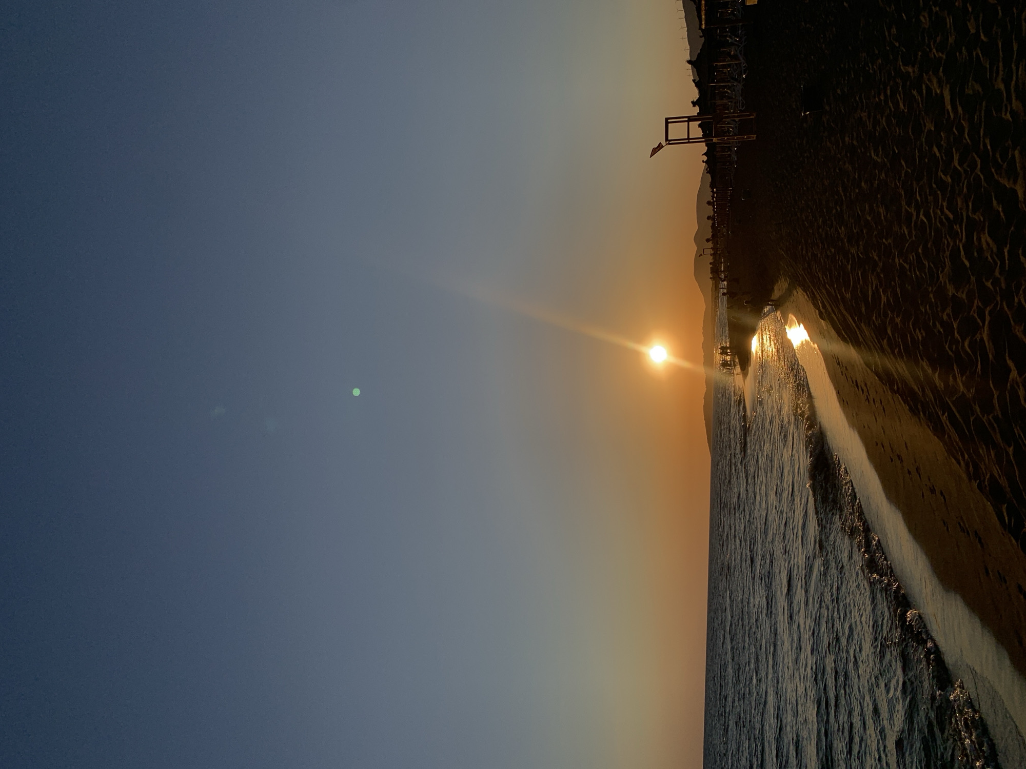 At sunset on a beach in Donji Štoj - Shtoji i Poshtëm, Montenegro, the sun is a bright orb low on the horizon, casting a warm orange glow across the sky and reflecting on the gentle waves of the Adriatic Sea. The foreground is dominated by a sandy beach with a few scattered footprints. In the mid-ground, the shoreline meets the water, with small waves lapping at the sand. Silhouettes of people are visible walking along the water's edge and on the beach. Further back, beach umbrellas and chairs are set up, indicating a developed tourist area. A lifeguard tower with a red flag is also visible. In the distance, hills or mountains form a dark silhouette against the vibrant sky. The upper portion of the image shows a gradient of clear blue sky transitioning to the orange hues of the sunset. There are two notable lens flares in the sky, one larger and more prominent near the center, and a smaller, fainter green one higher up.