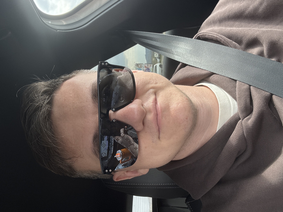A male individual with short brown hair, wearing dark, rectangular sunglasses, a brown hooded sweatshirt over a white t-shirt, and a dark grey seatbelt, is seated in the front passenger or driver's seat of a vehicle. The individual's expression shows a slight smile. Reflections in the sunglasses reveal an orange insulated tumbler with a straw and a light-colored item featuring a repeating Louis Vuitton monogram pattern, both situated within the vehicle's interior. Through the vehicle's window and in the reflections, a road or highway environment is visible, featuring a distant white van, a large billboard with multiple panels, and hills or mountains under a cloudy sky. The vehicle's interior is dark-toned. The scene is located in Pasohlávky, Czechia.