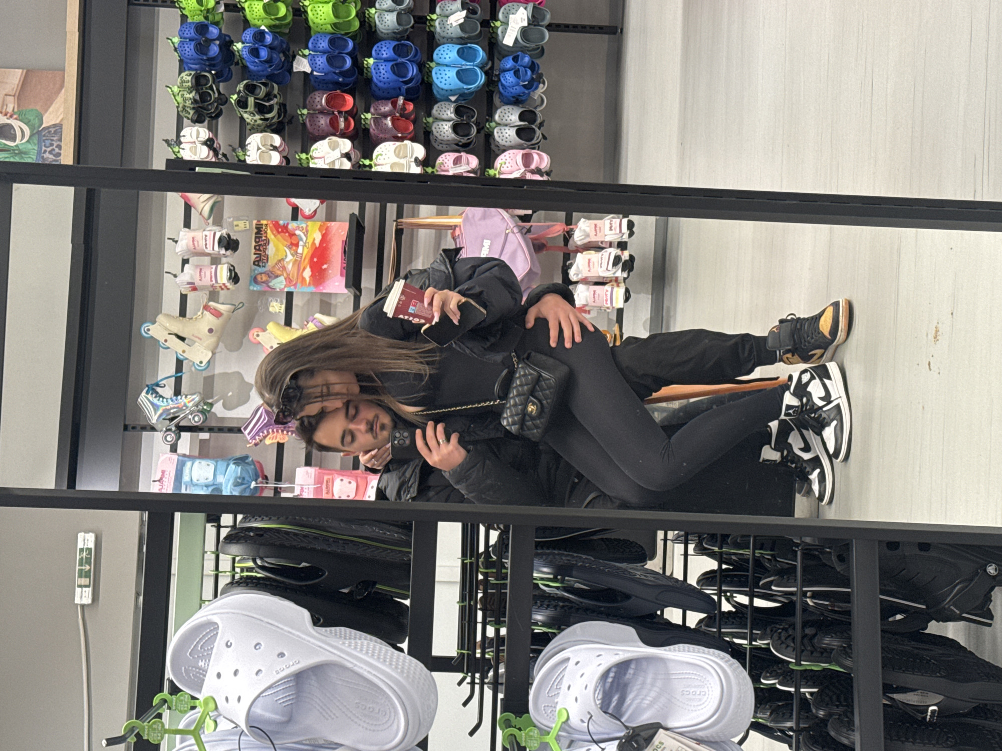 The image displays a reflection of a couple embracing in what appears to be a retail store. The woman, with long brown hair, wears a black jacket, black leggings, and black and white high-top sneakers. Sunglasses are pushed up onto her head, and she carries a black quilted shoulder bag. She is holding a book or passport and a smartphone. The man, with a beard, wears a dark jacket and pants, and also holds a smartphone, with his arm around the woman. Their affectionate embrace suggests a personal moment captured within a public space.
The setting is an indoor store, characterized by numerous shelves displaying footwear and accessories. Prominently featured are multiple pairs of Crocs in various colors including green, blue, grey, maroon, pink, white, and black, arranged on black shelving units. White Crocs are visible in the lower foreground. Other merchandise includes rollerblades, small bags (one purple), and packaged socks. A framed picture depicting an astronaut-like figure hangs on the wall to the far left. The word "ALADDIN" is visible on a product box in the background, and the brand name "CROCS" is discernible on some of the clogs. The scene suggests a daytime visit to a casual shopping environment.