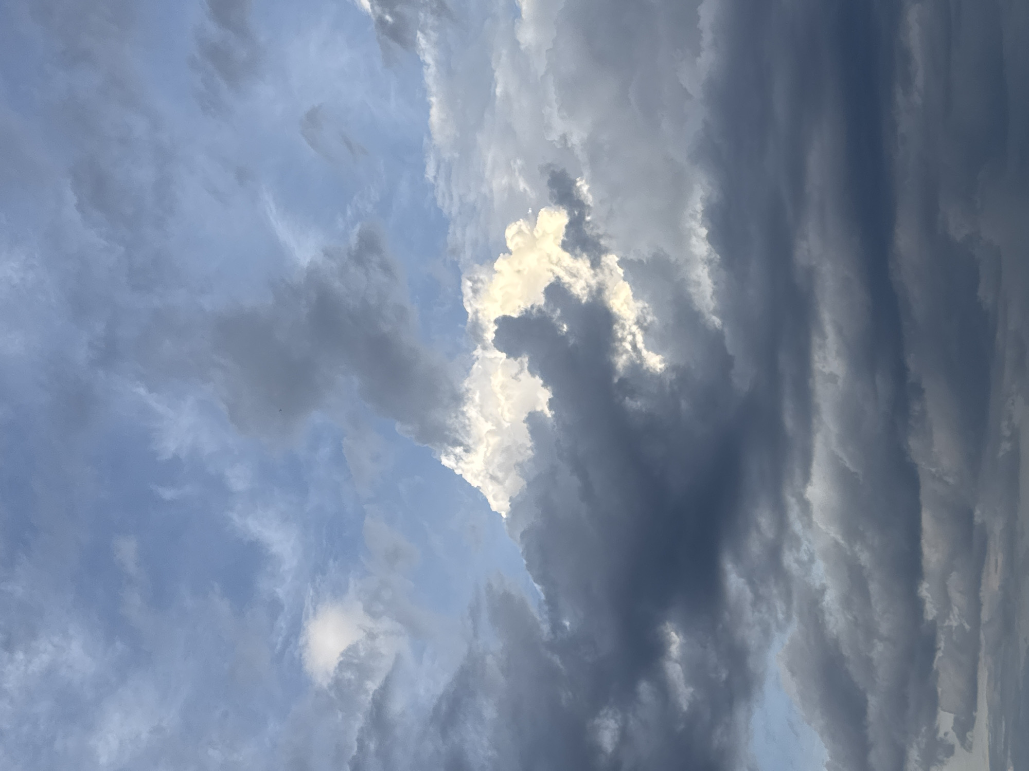 The image shows a dramatic sky filled with a variety of clouds. In the center, a large, bright white cloud is illuminated by the sun, with darker, more ominous clouds surrounding it. The sky transitions from a pale blue in the upper portions to darker grays and blues towards the bottom, suggesting an impending storm or the aftermath of one. There are no people, subjects, or objects visible in the image, and no actions or interactions are depicted. The scene is entirely focused on the atmospheric conditions. The location is identified as Nha Trang, Vietnam, implying this is a view of the sky above the city.