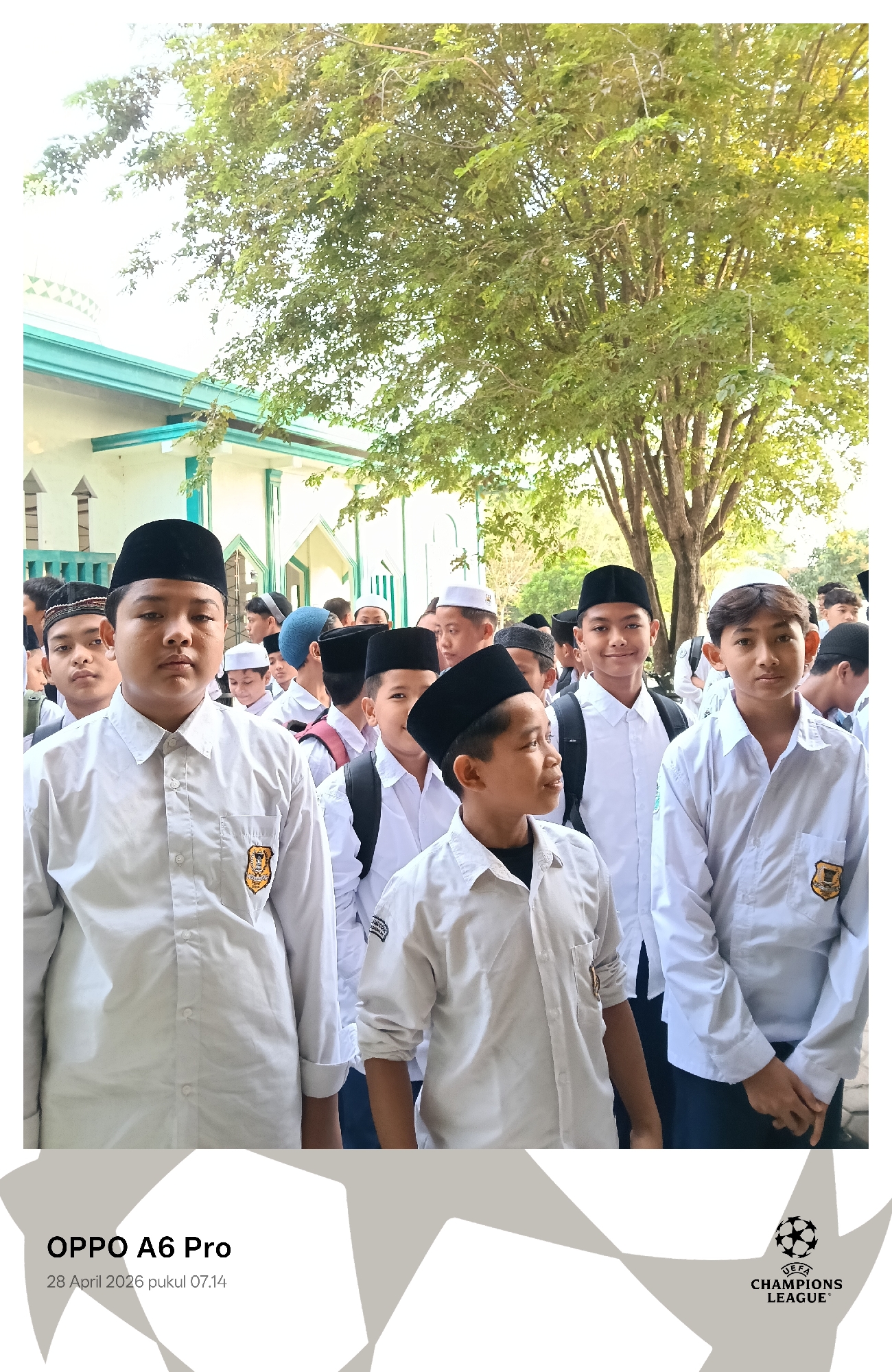 The image displays a group of young boys, likely students, gathered outdoors in Lhokseumawe, Indonesia, on the morning of April 28, 2026, at 07:14 AM. The scene is bathed in bright daylight under a clear sky.
The main subjects are the boys, who appear to be between elementary and middle school age. Most are dressed in matching white, long-sleeved shirts and wear traditional peci (skullcaps) – some black, others white – suggesting a uniform or attire for an Islamic educational or religious setting. Several boys carry backpacks, further indicating they might be students attending school.
In the foreground, a boy wearing a black peci and a white shirt with a yellow and black emblem on his pocket stands prominently, looking directly at the camera. To his left, another boy in a similar uniform looks off to the side, while to his right, a boy with a black peci gazes rightward. On the far right, a boy in a white peci looks towards the camera. The boys generally exhibit calm expressions, some looking at the camera, others engaged with their surroundings or each other.
The background features a light-colored building with distinctive green trim and a partial dome-like structure visible on the left, strongly suggesting a mosque or an Islamic school (madrasah). Behind the building and the boys, a large, mature tree with lush green foliage and visible branches provides a natural and shaded backdrop.
The activity appears to be a gathering or assembly, possibly before classes or a religious event.
Notable details include the uniform-like attire of the boys, the architectural cues of the building, and the bright morning light. Visible text watermarks include "OPPO A6 Pro," the date "28 April 2026 pukul 07.14," and a "UEFA CHAMPIONS LEAGUE" logo.