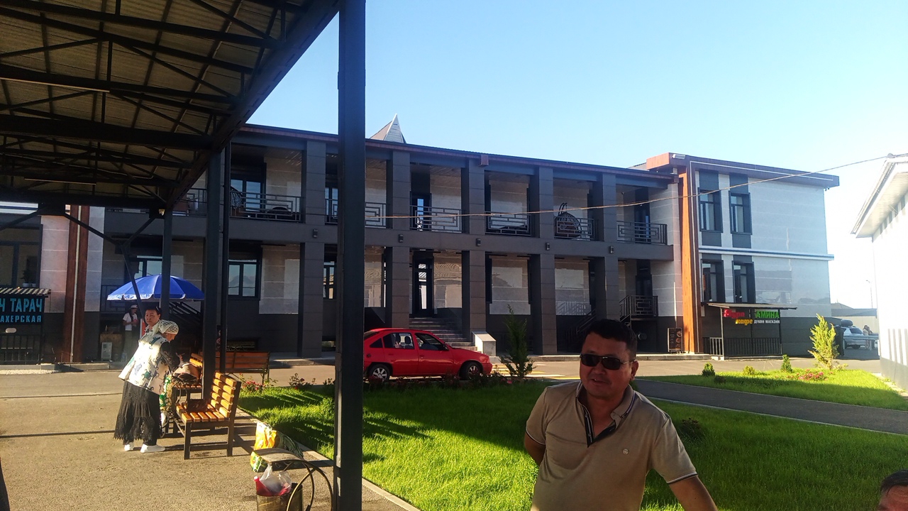 In Balykchy, Kyrgyzstan, on a clear day, a man wearing a light brown polo shirt and sunglasses stands in the foreground, facing right. Behind him, a multi-story building with a modern facade, featuring dark columns and balconies, is visible. A red car is parked on the street in front of the building. To the left, two women are standing near a blue umbrella and a wooden bench. The scene is set outdoors with a patch of green grass and a paved area in front of the building. The overall impression is of a commercial or residential area on a sunny day.