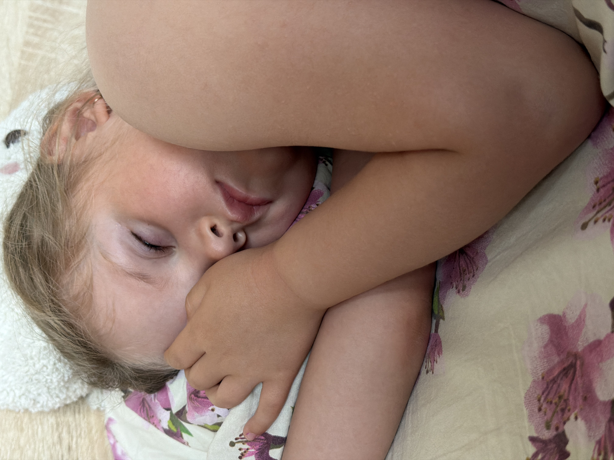 A close-up, high-angle shot captures a young child peacefully sleeping or resting. The child, with fair skin and light blonde or light brown hair, is lying down with their eyes closed and one arm partially covering their face. Their features appear soft and relaxed, conveying an emotion of tranquility. A small, golden earring is visible in the child's ear.
The child's head rests on a white terrycloth towel or blanket, which has a fluffy texture and a subtle animal-like ear shape visible on the left side, possibly part of a hooded towel or blanket. The child is partially covered by or lying on a floral patterned fabric, likely bedding, which features large pink and purple flowers with green leaves on a light, cream-colored background. The setting appears to be indoors, possibly a bedroom. There are no direct cues about the time of day or weather, nor any visible text. The provided location context is Chaikovsky, Russia.